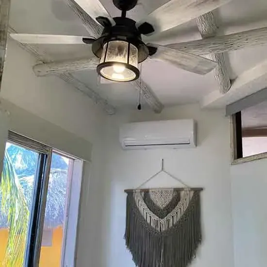 Ventilador de techo con luz, vigas encaladas, arte de pared, unidad de aire acondicionado y ventana en una habitación.
