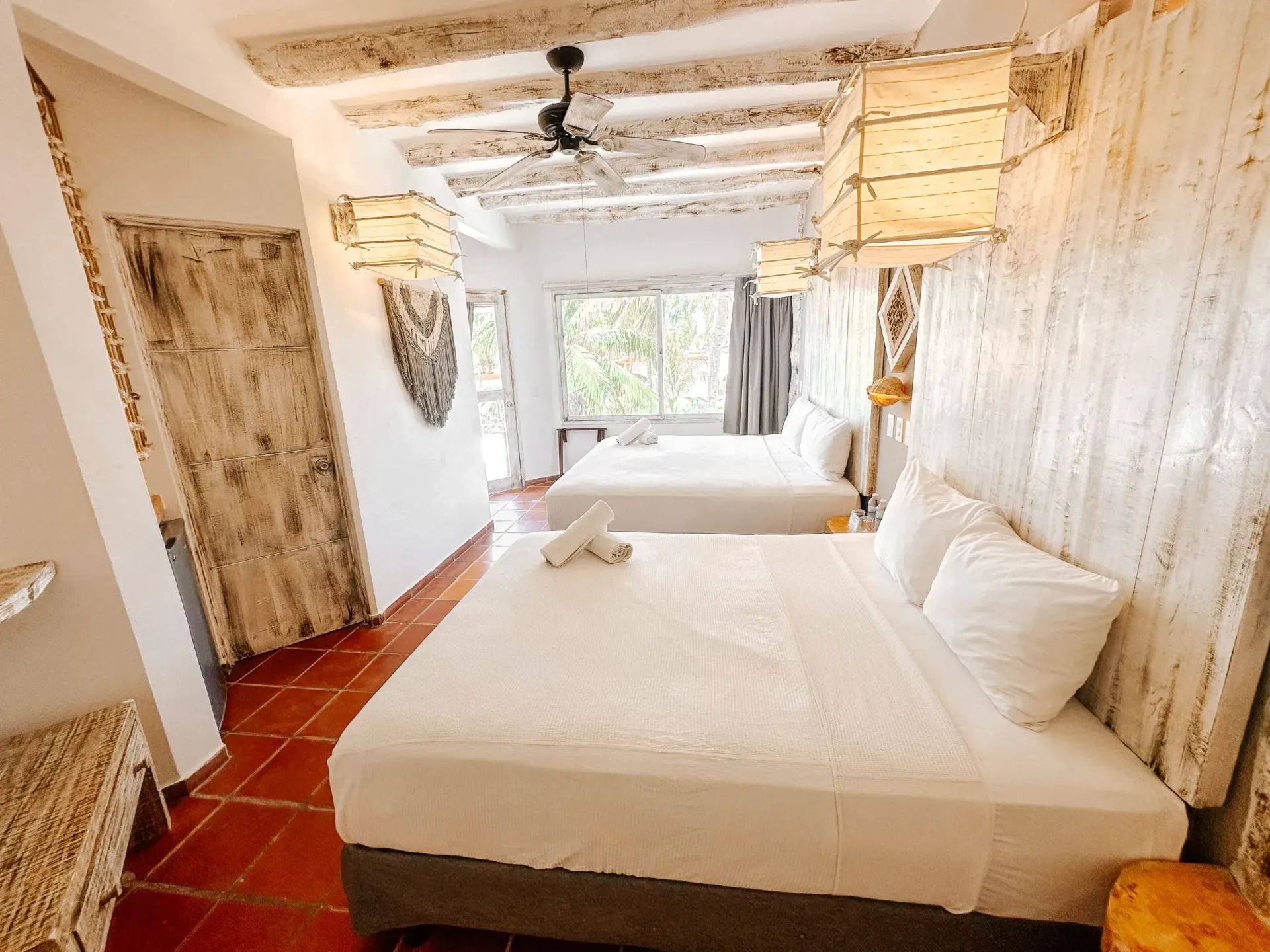 Dormitorio con dos camas, paredes blancas y lámparas colgantes decorativas. Puerta de madera y suelo de baldosas rojas.