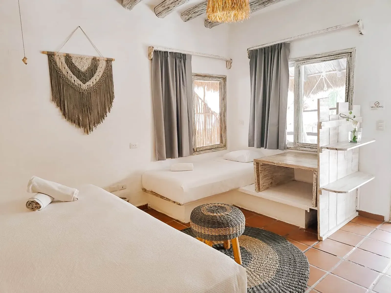 Dormitorio con dos camas, tonos neutros, tapiz de macramé, ventana con cortinas grises y escritorio.