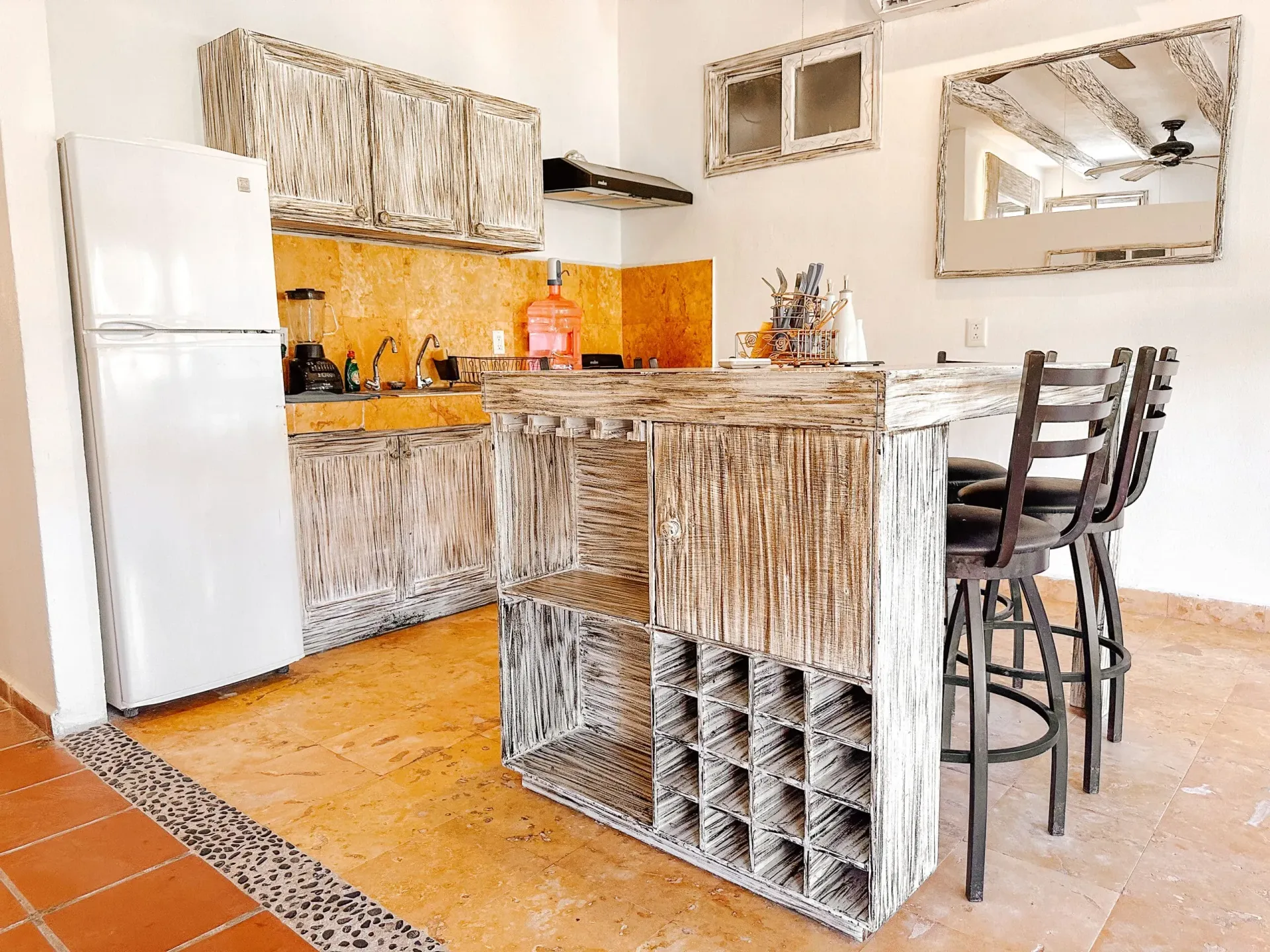 Cocina con refrigerador blanco, gabinetes símil madera y barra desayunadora, taburetes, piso de baldosas color naranja.