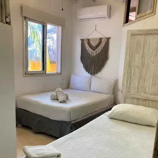 Dormitorio con dos camas dobles, paredes blancas, ventana, tapiz de macramé y aire acondicionado.