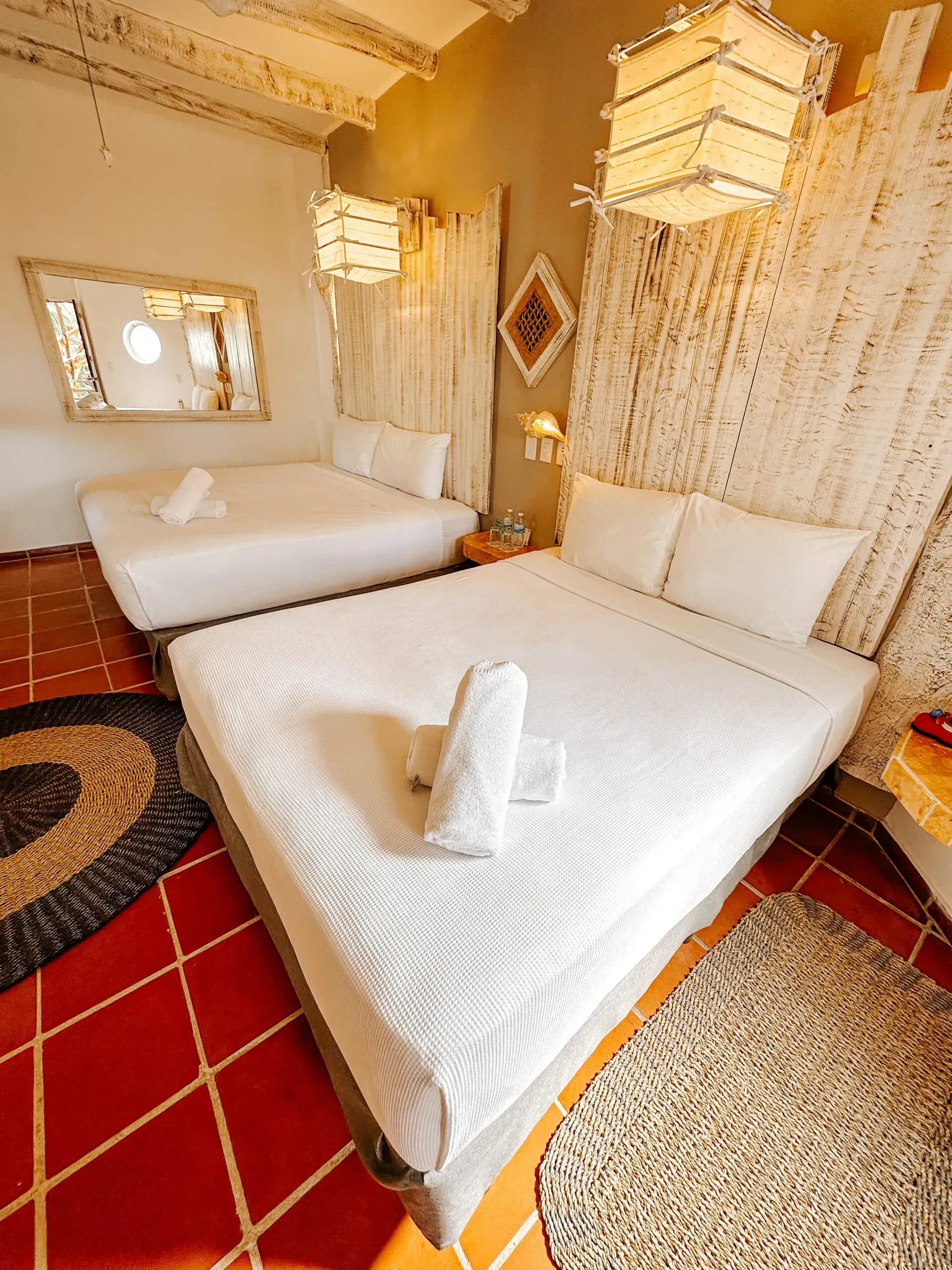 Acogedora habitación de hotel con dos camas blancas, suelo de baldosas rojas y alfombras tejidas. Lámparas decorativas.