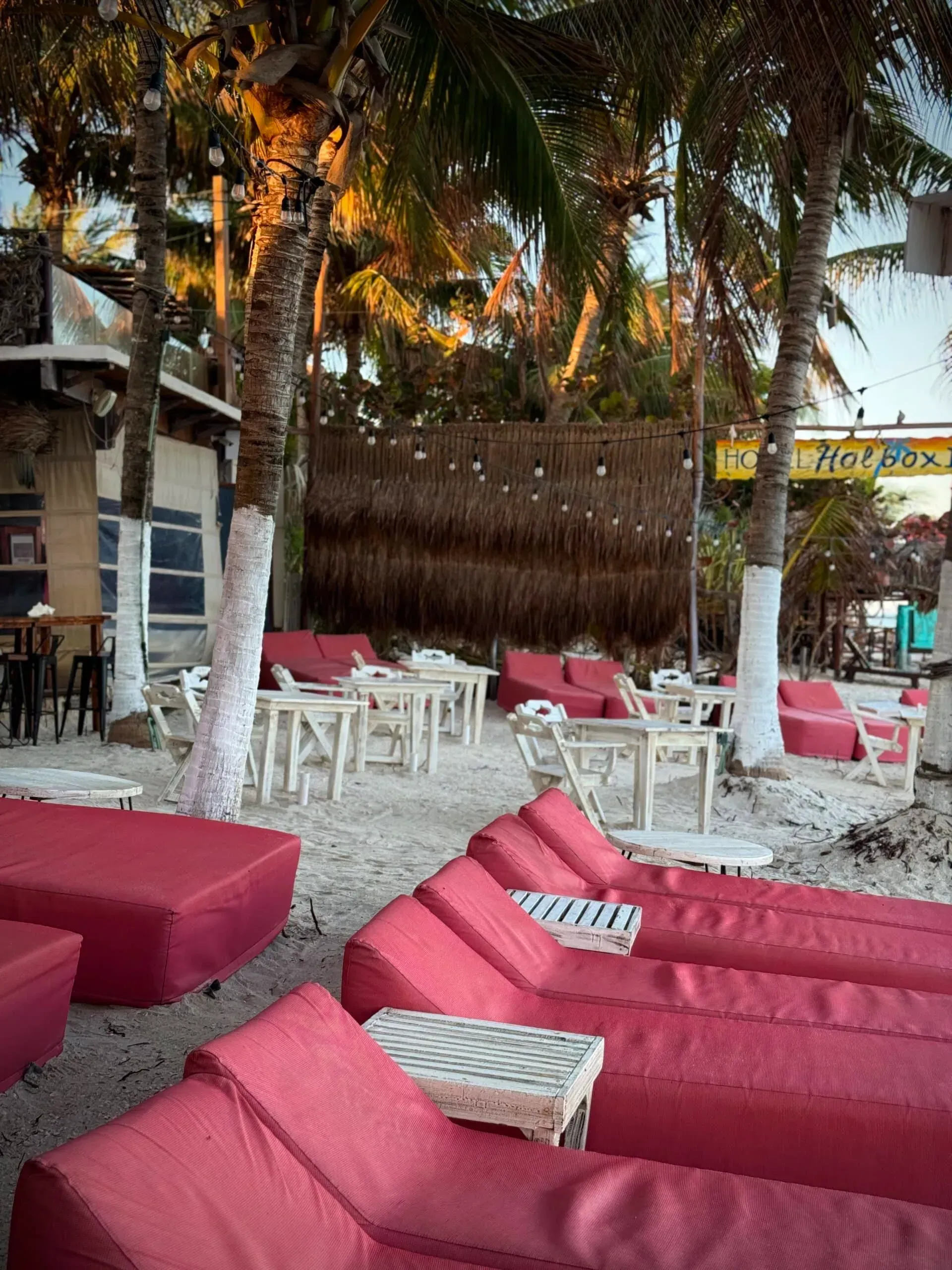 Salón junto a la playa con cojines rojos, mesas blancas y palmeras, bañado por la cálida luz del sol.