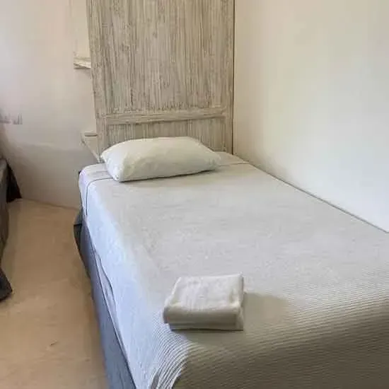 Una cama individual con cabecera blanca, almohada y toalla doblada; paredes blancas.