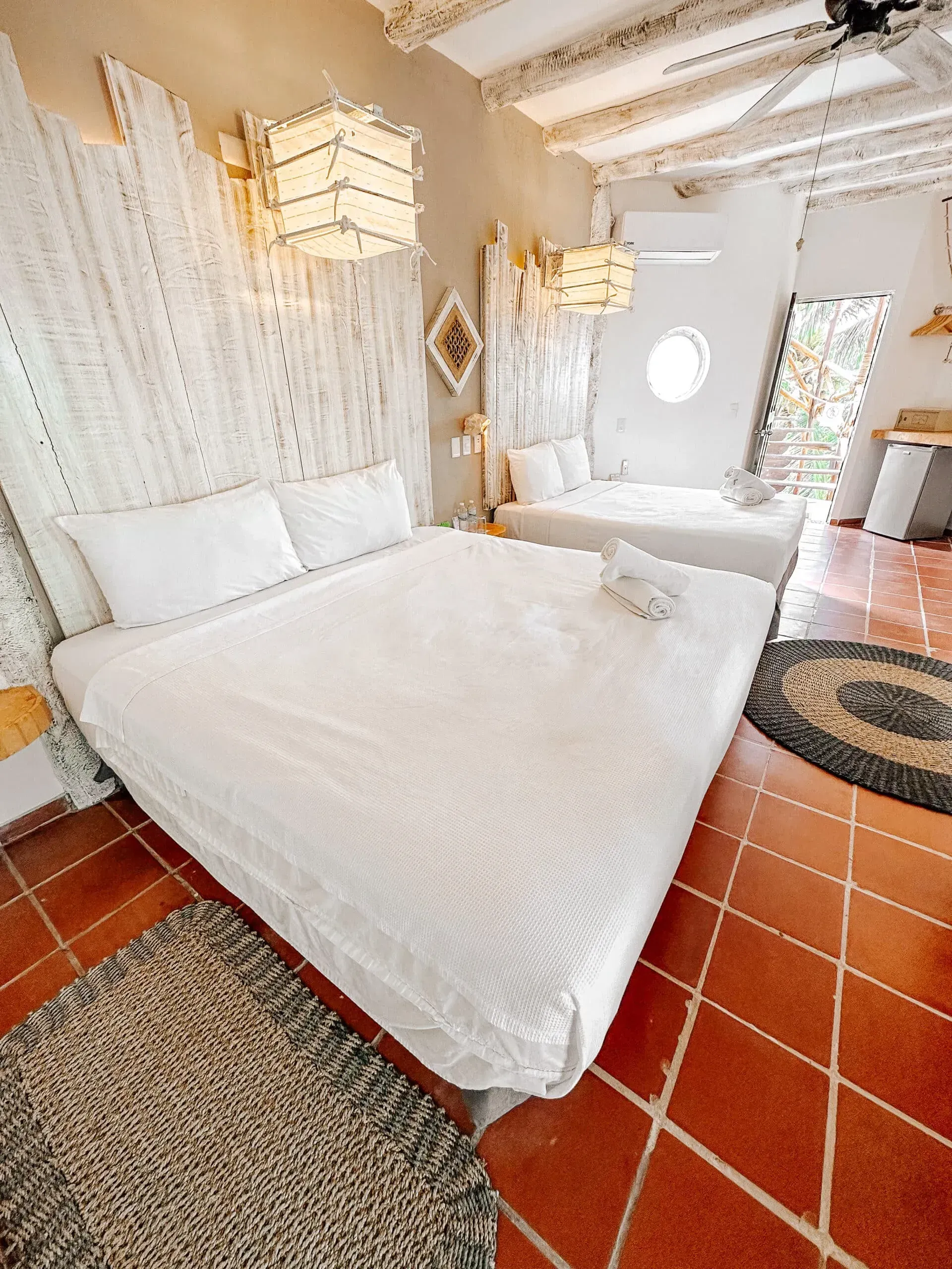 Dormitorio con dos camas blancas, cabeceros tejidos en color blanco y suelo de baldosas de terracota.