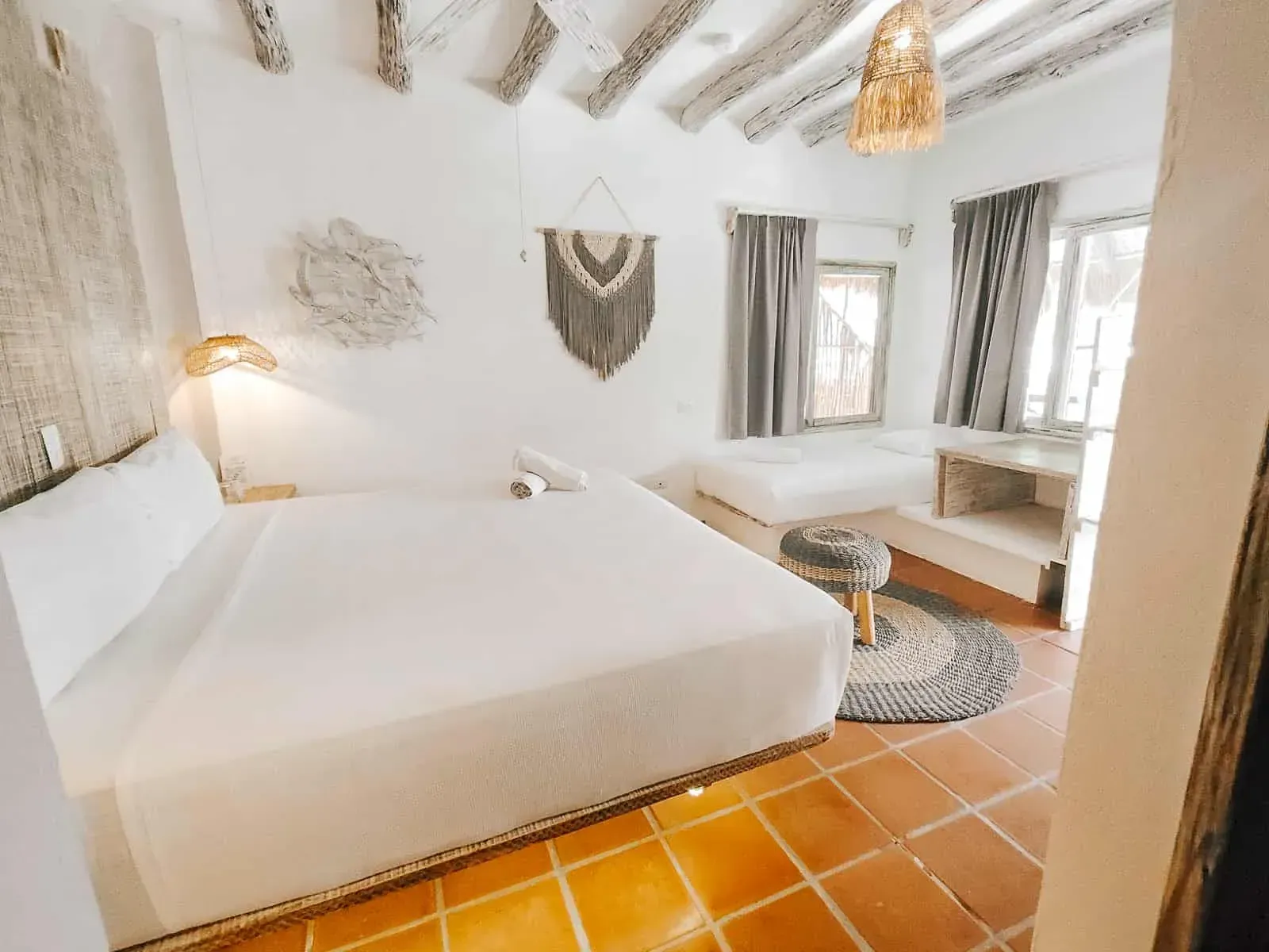 Habitación de hotel de color blanco brillante con cama, zona de estar, detalles tejidos y piso de baldosas de terracota.