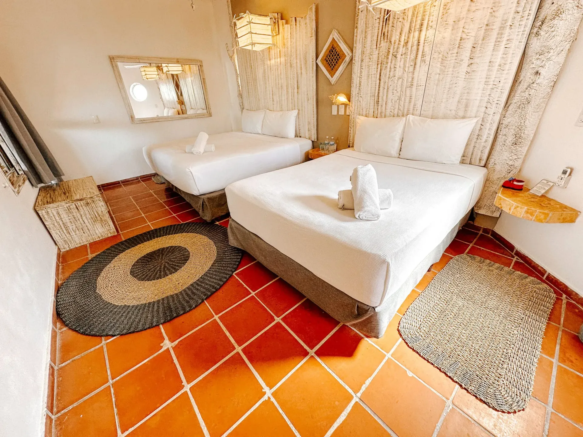 Acogedora habitación de hotel con dos camas, ropa de cama blanca, alfombras tejidas y suelo de baldosas de terracota.
