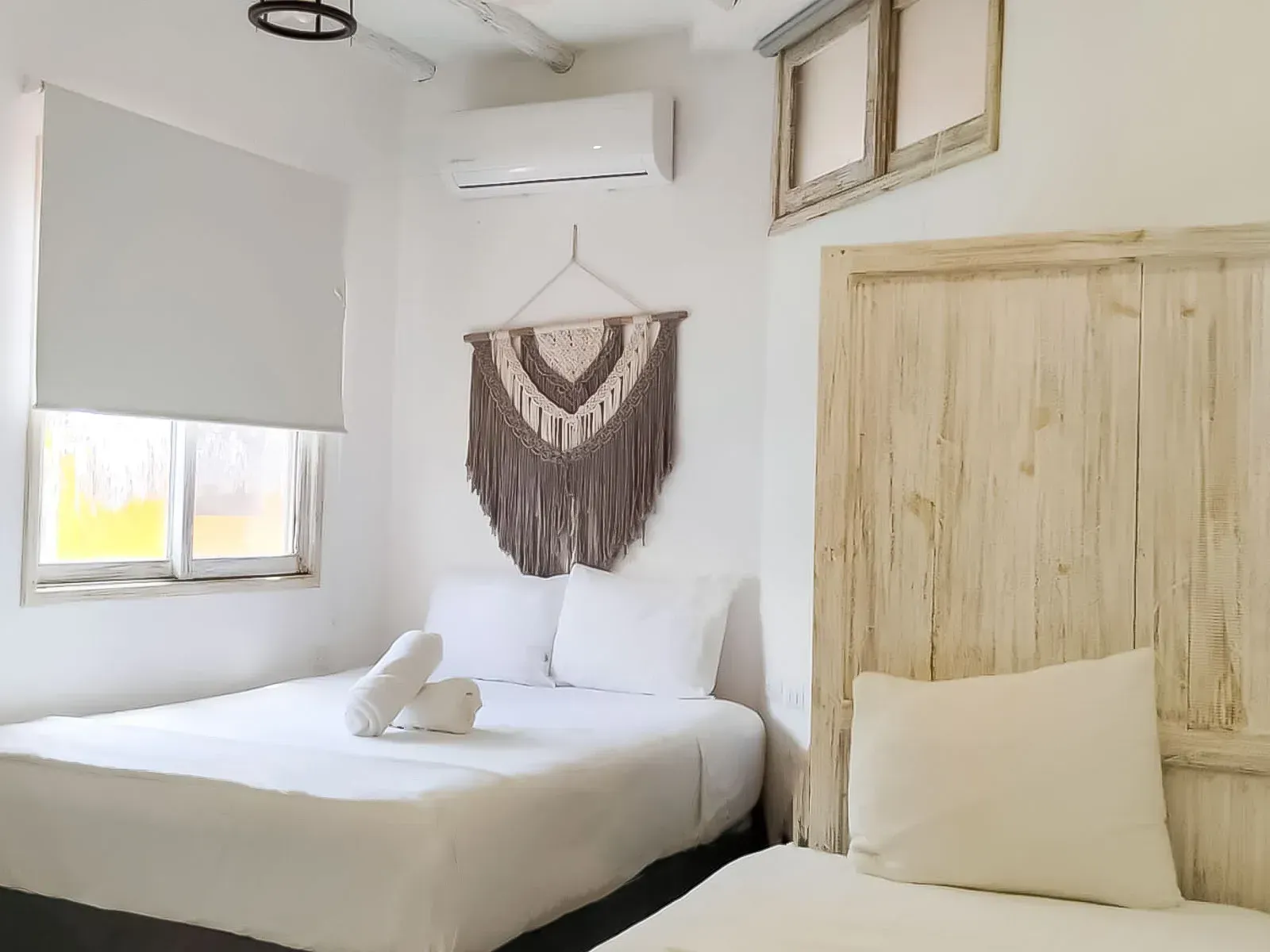 Dormitorio con ropa de cama blanca, tapiz de macramé y unidad de aire acondicionado.