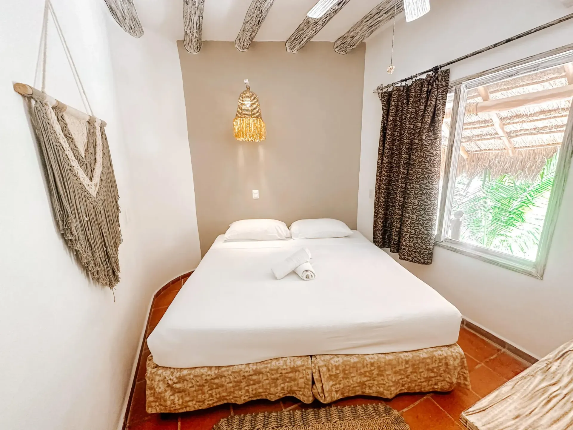 Acogedor dormitorio con cama blanca, tapiz de macramé y pequeña ventana con vista verde.