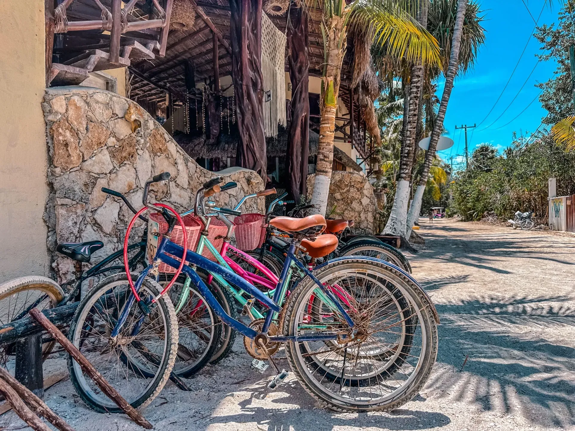 Bicicletas estacionadas junto a un edificio con una pared de piedra texturizada, palmeras y un cielo soleado.