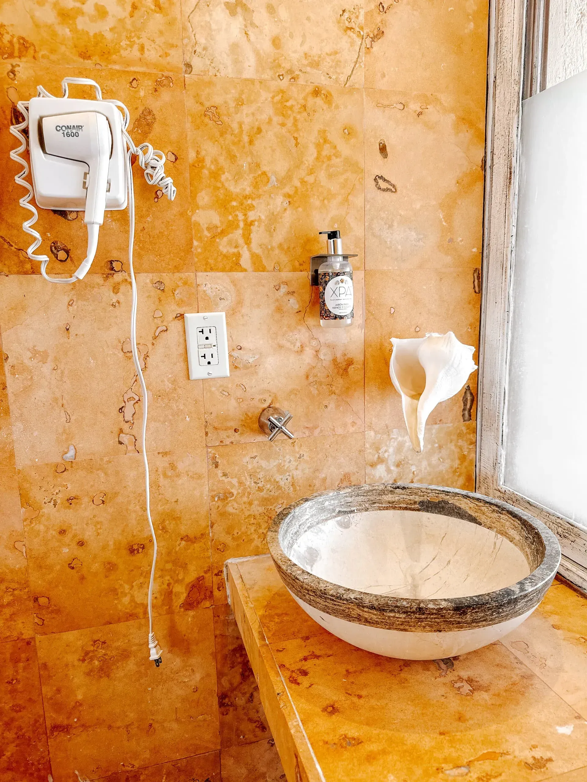 Baño con pared de piedra naranja, lavabo con forma de concha, secador de pelo, dispensador de jabón.