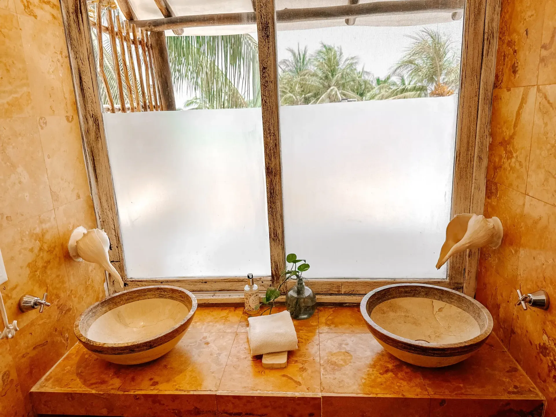 Baño con dos lavabos beige bajo una ventana esmerilada, con grifería en forma de concha y paredes de piedra.