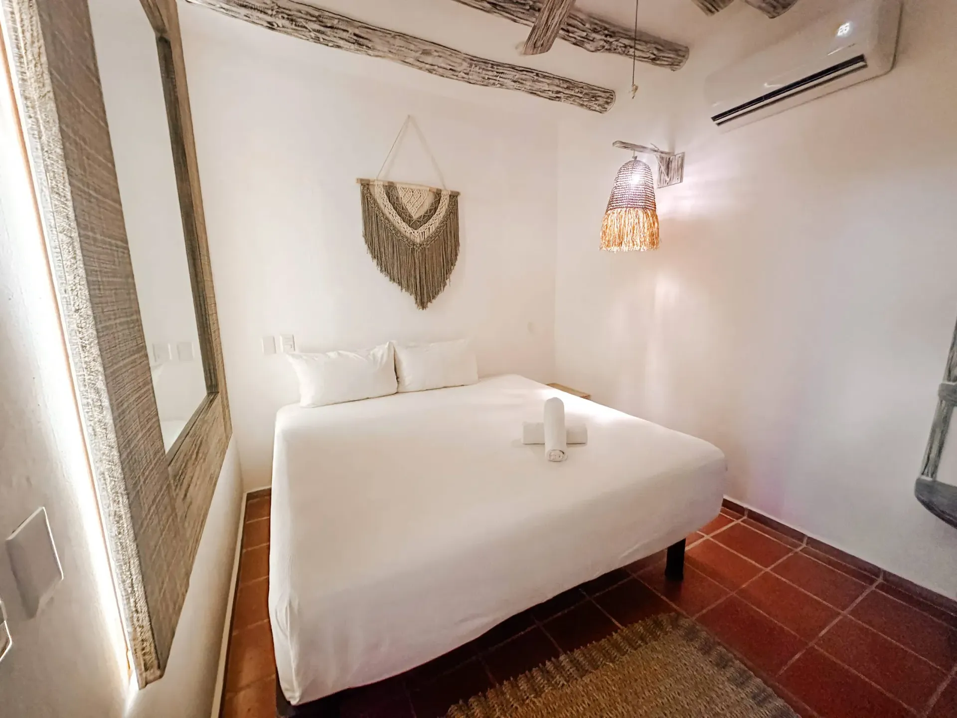 Pequeño dormitorio blanco con cama, tapiz de macramé, espejo y aire acondicionado.