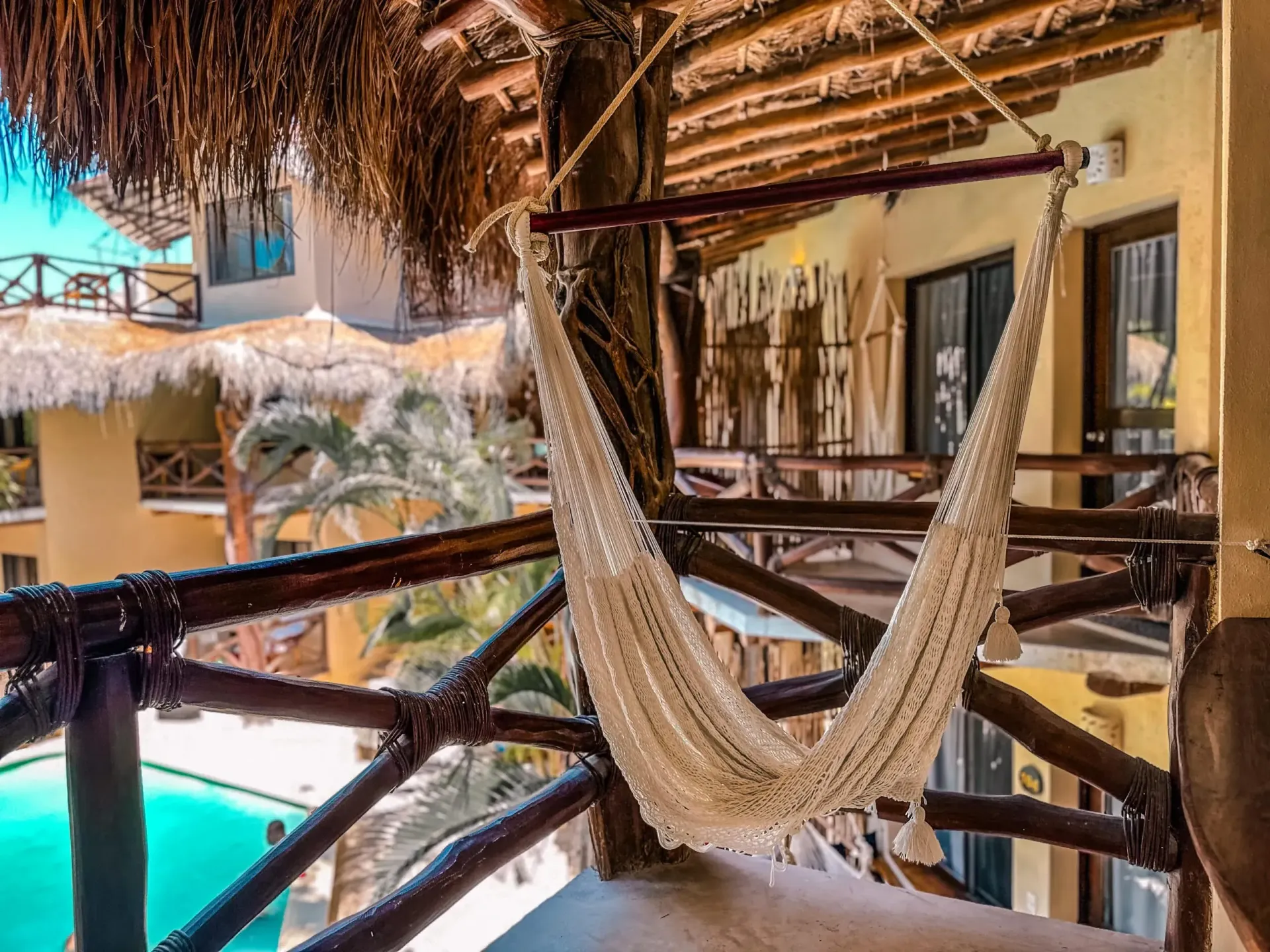 Una hamaca cuelga de un balcón con vista a una piscina en un resort tropical, con techo de paja y palmeras.