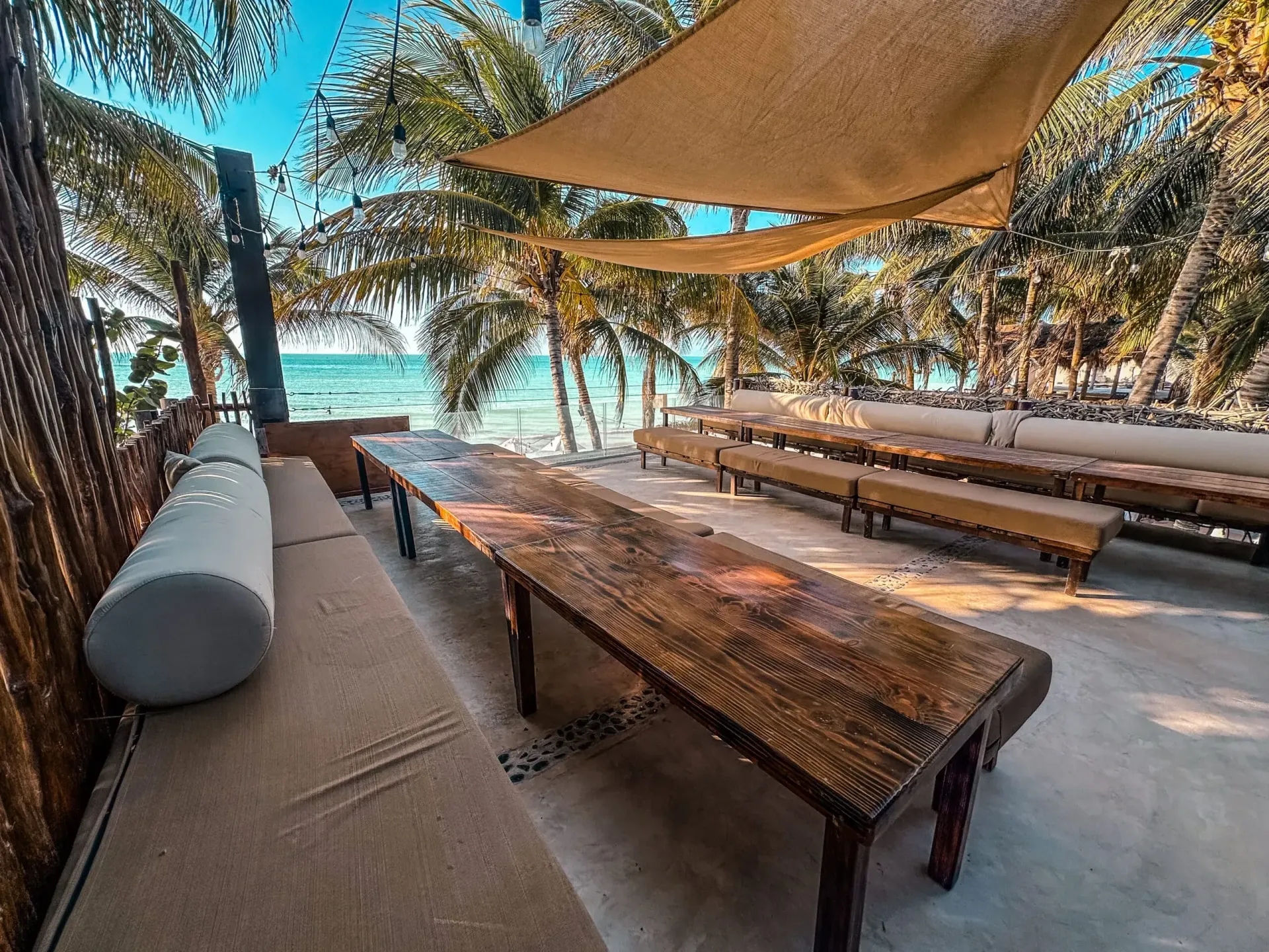 Zona de estar junto a la playa con largas mesas de madera, bancos acolchados y toldos para dar sombra; océano de fondo.