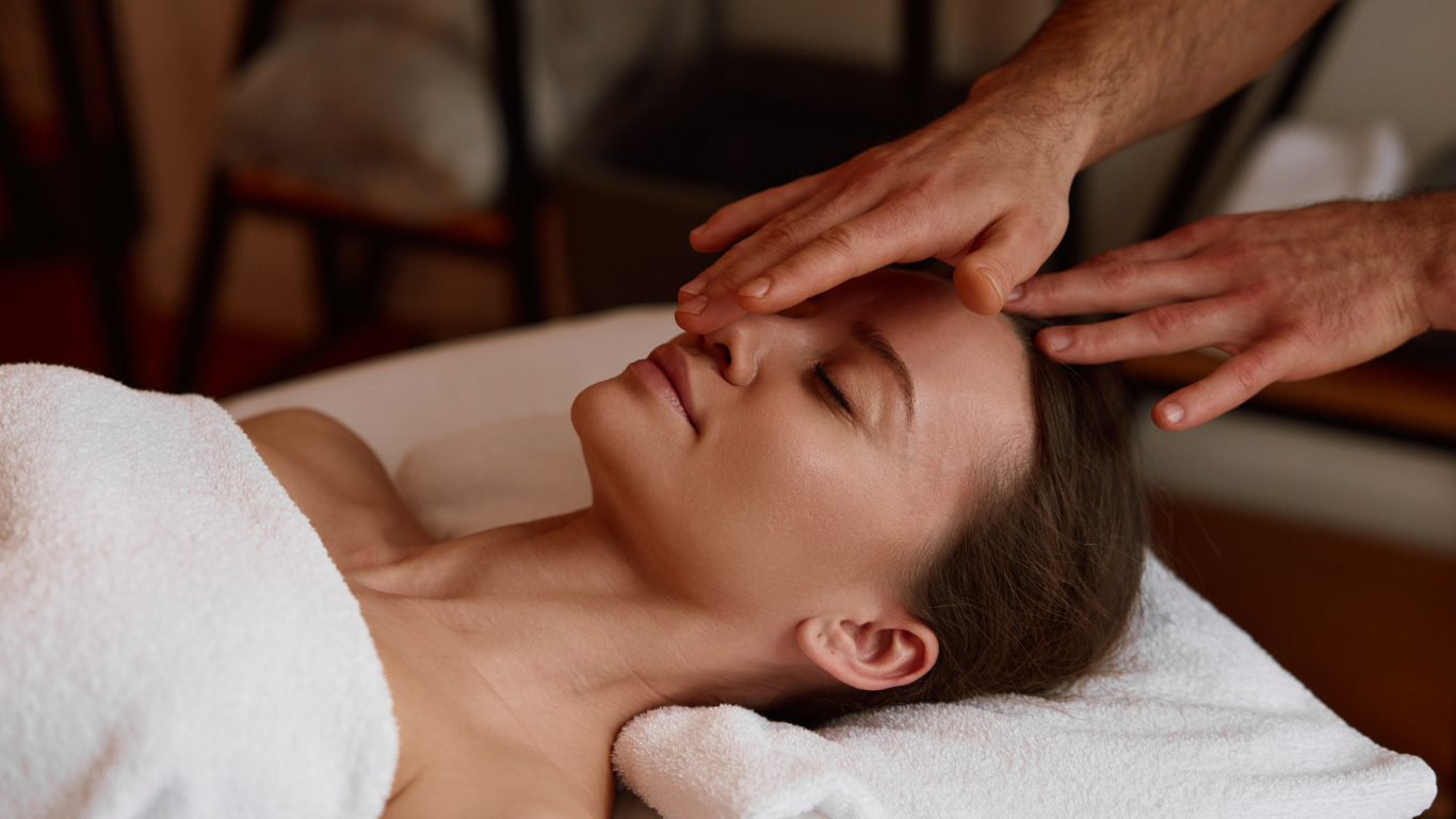 Person receiving head massage, eyes closed, hands on forehead, lying on massage table.