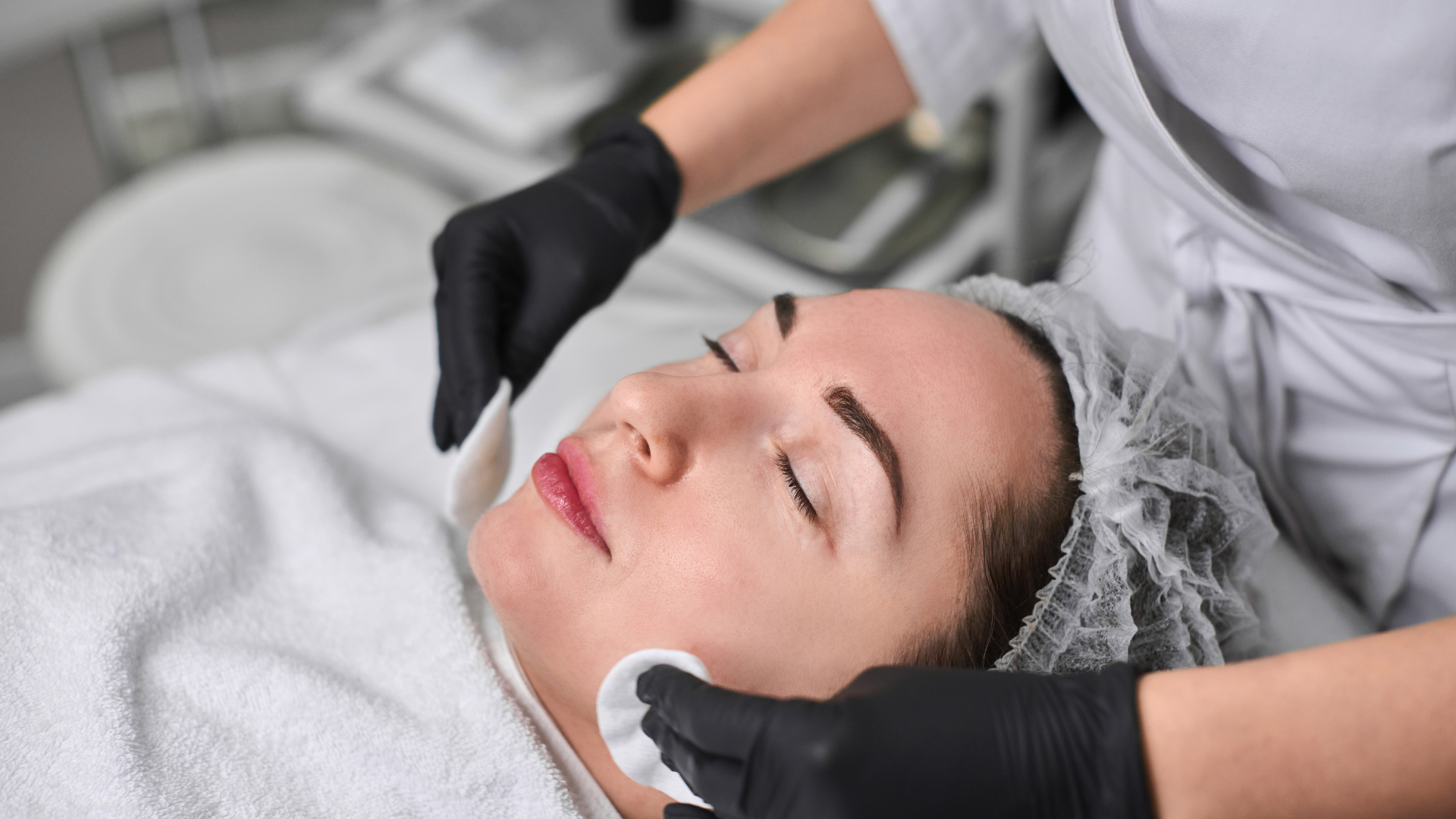 Person receiving a facial treatment; gloved hands cleansing face with cotton pads, spa setting.