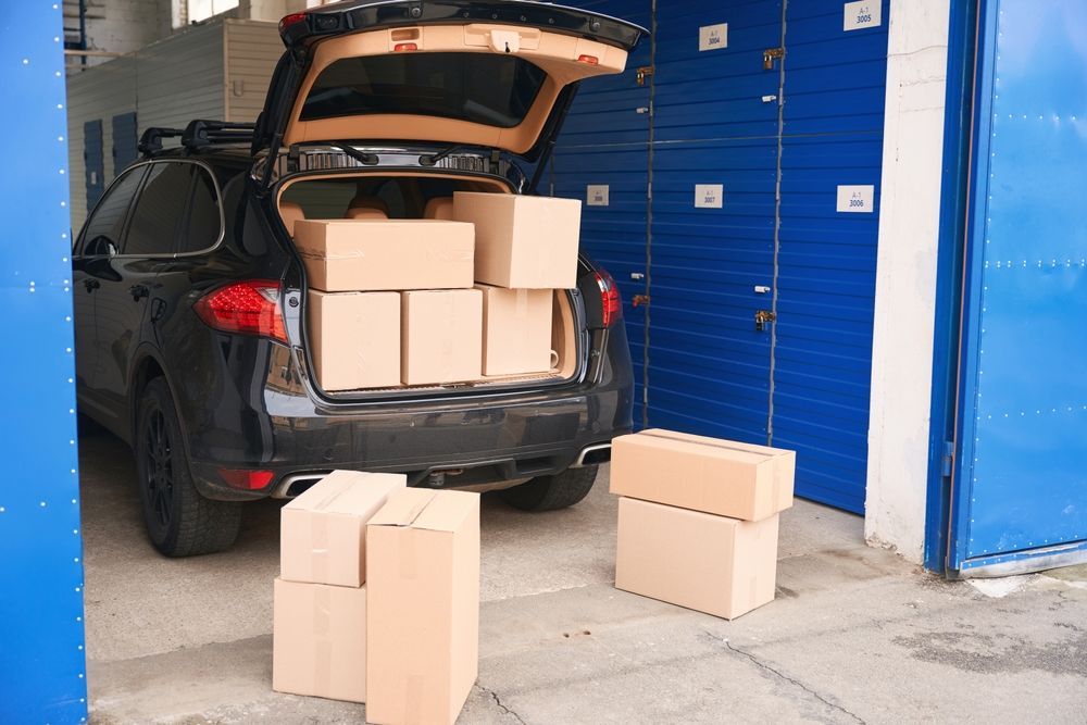 Black SUV with open trunk, filled with moving boxes, parked by blue storage units.