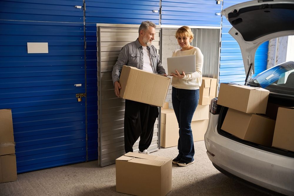 Couple loading cardboard boxes into a car at a storage unit, one holds a laptop.