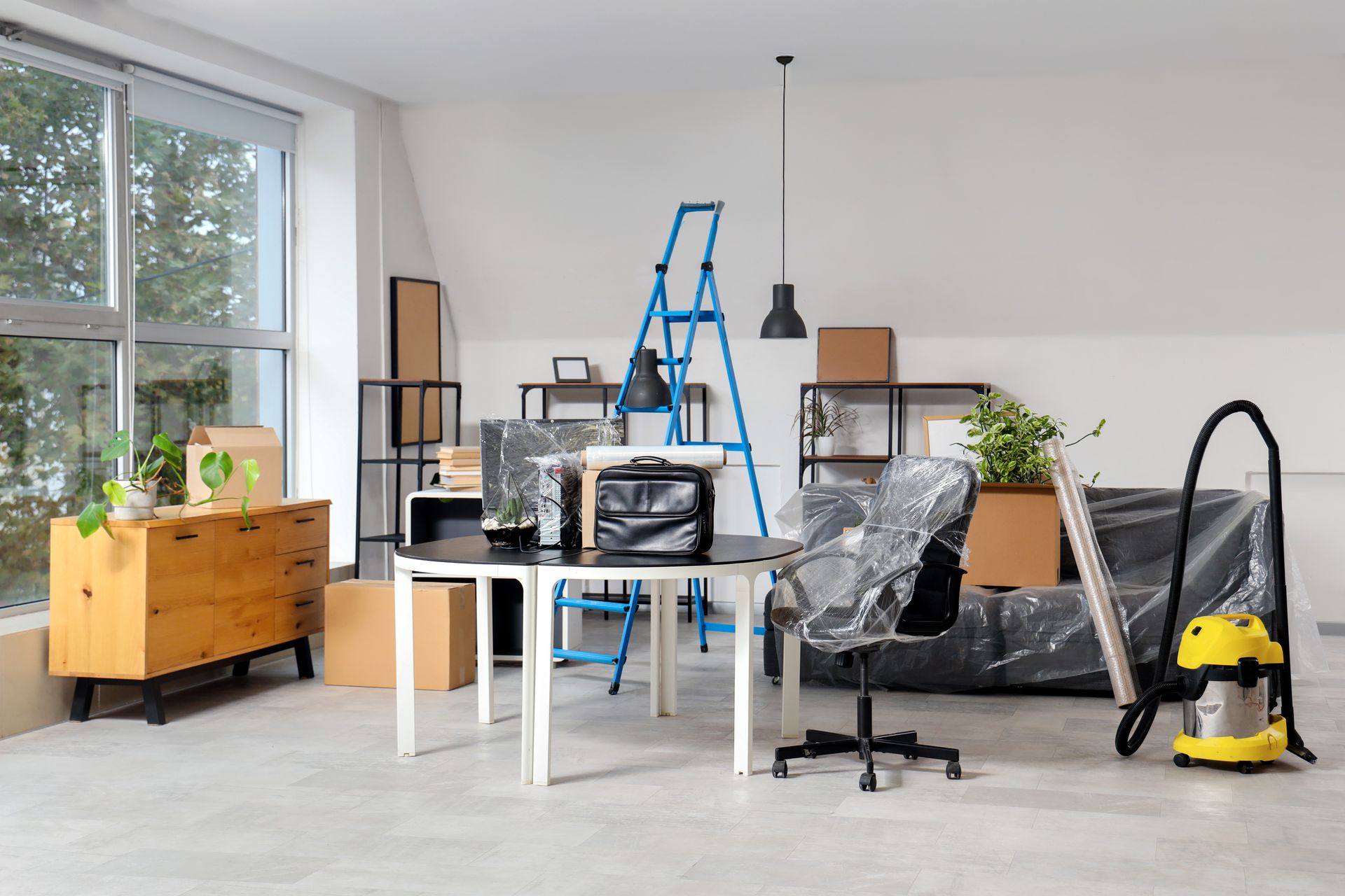 Office interior during a move. Packed boxes, furniture covered in plastic, ladder, and vacuum cleaner.