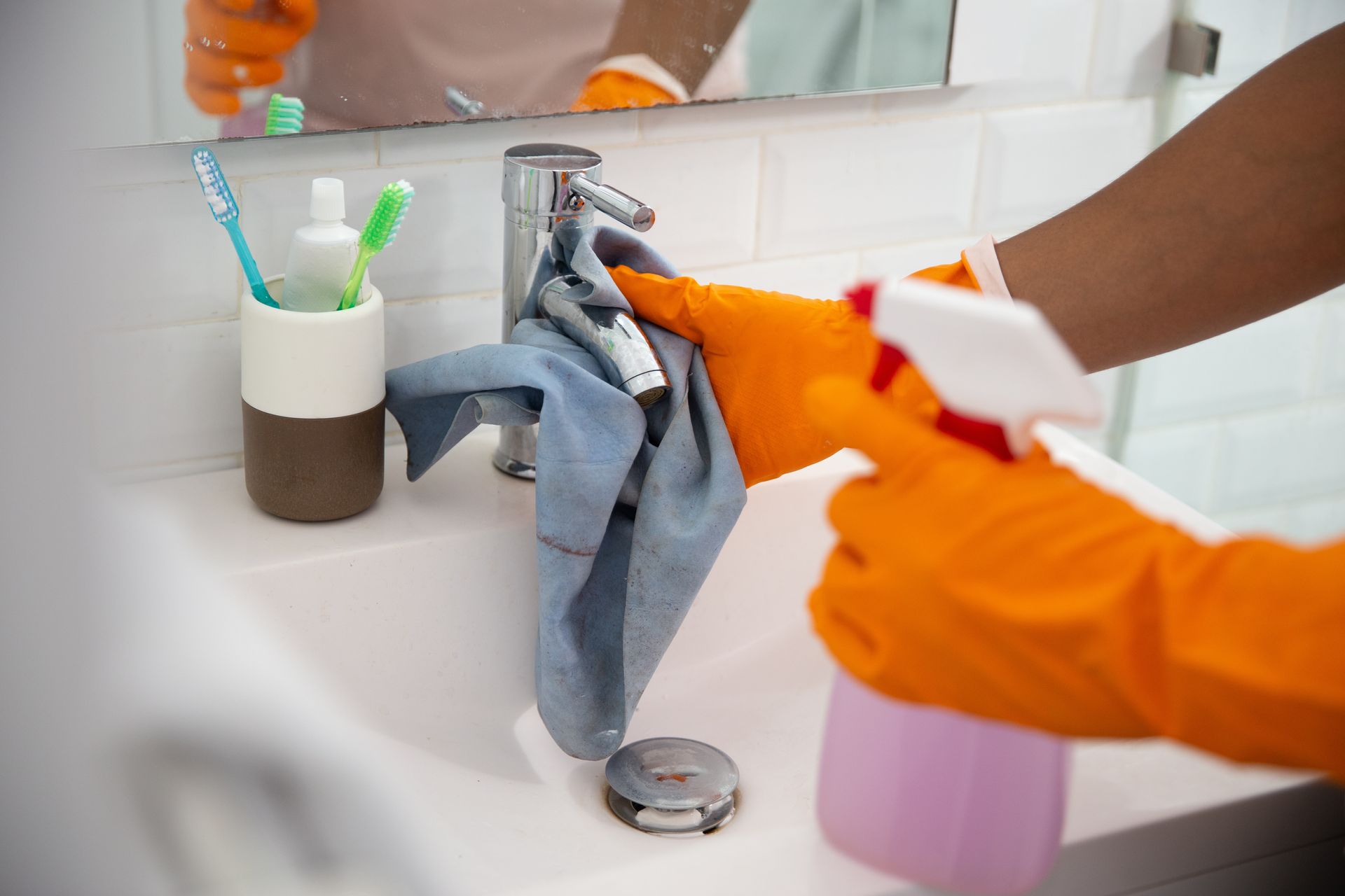 Person wearing orange gloves cleaning a bathroom sink with a spray bottle and cloth.