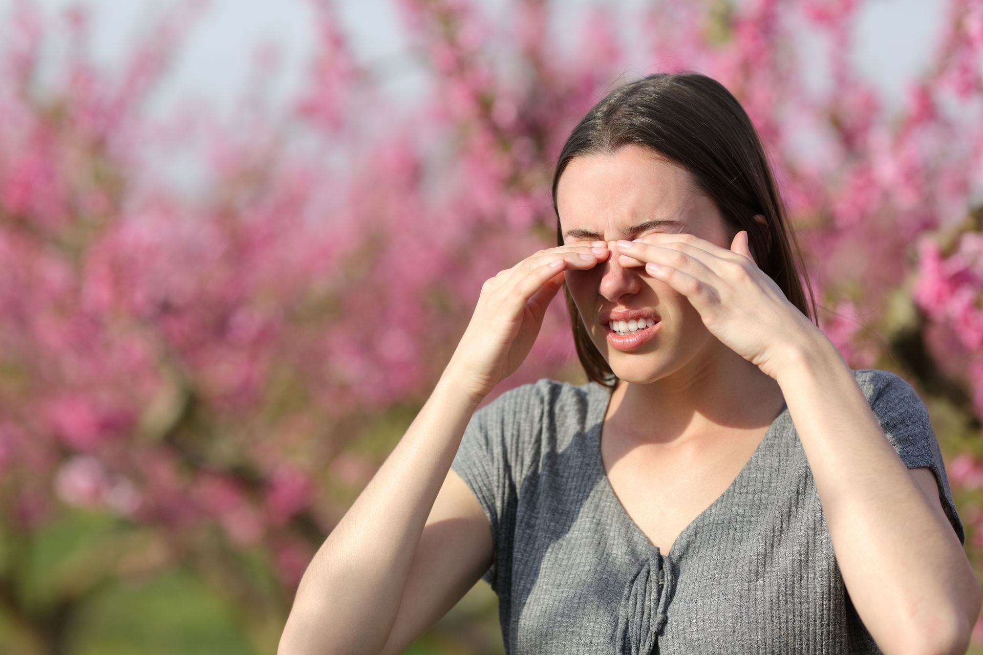 A woman is rubbing her eyes in front of a field of pink flowers.