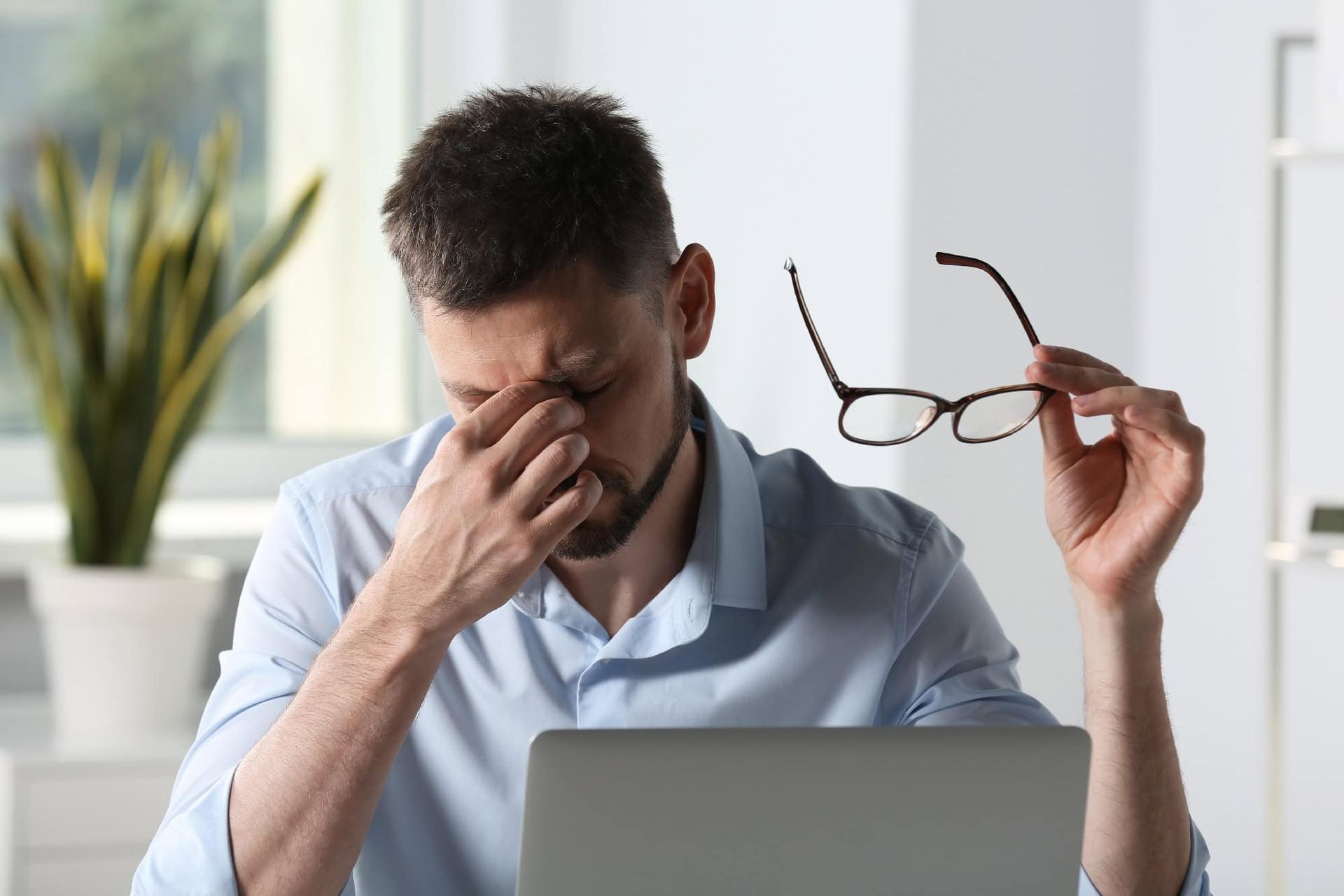 A man is sitting in front of a laptop computer holding his glasses.