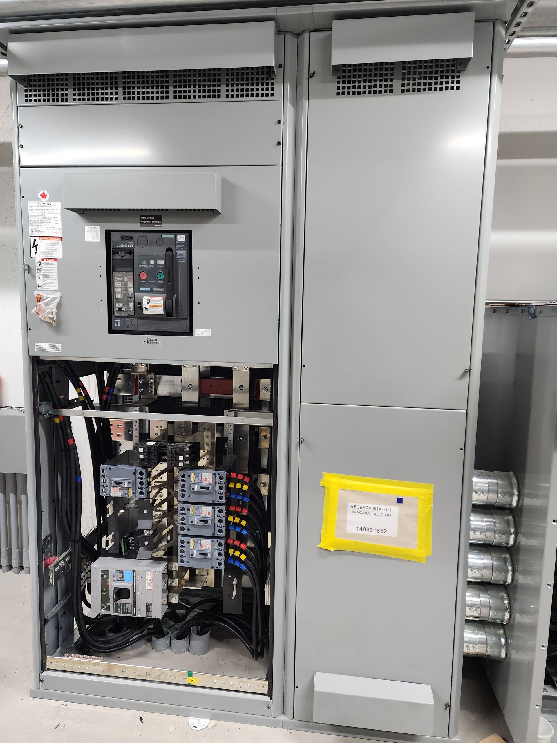 Electrical panel cabinet, gray, open on the left revealing wiring and components, with a large circuit breaker visible.