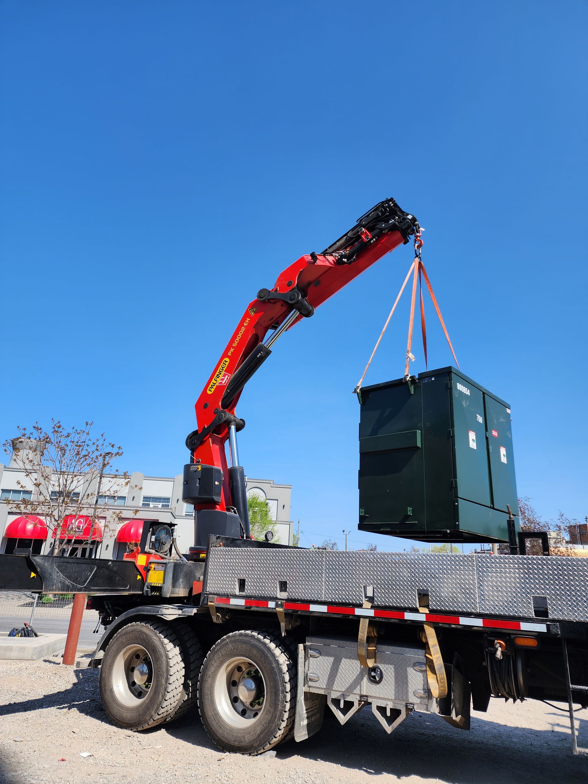 A red crane on a truck lifts a dark green box against a bright blue sky.
