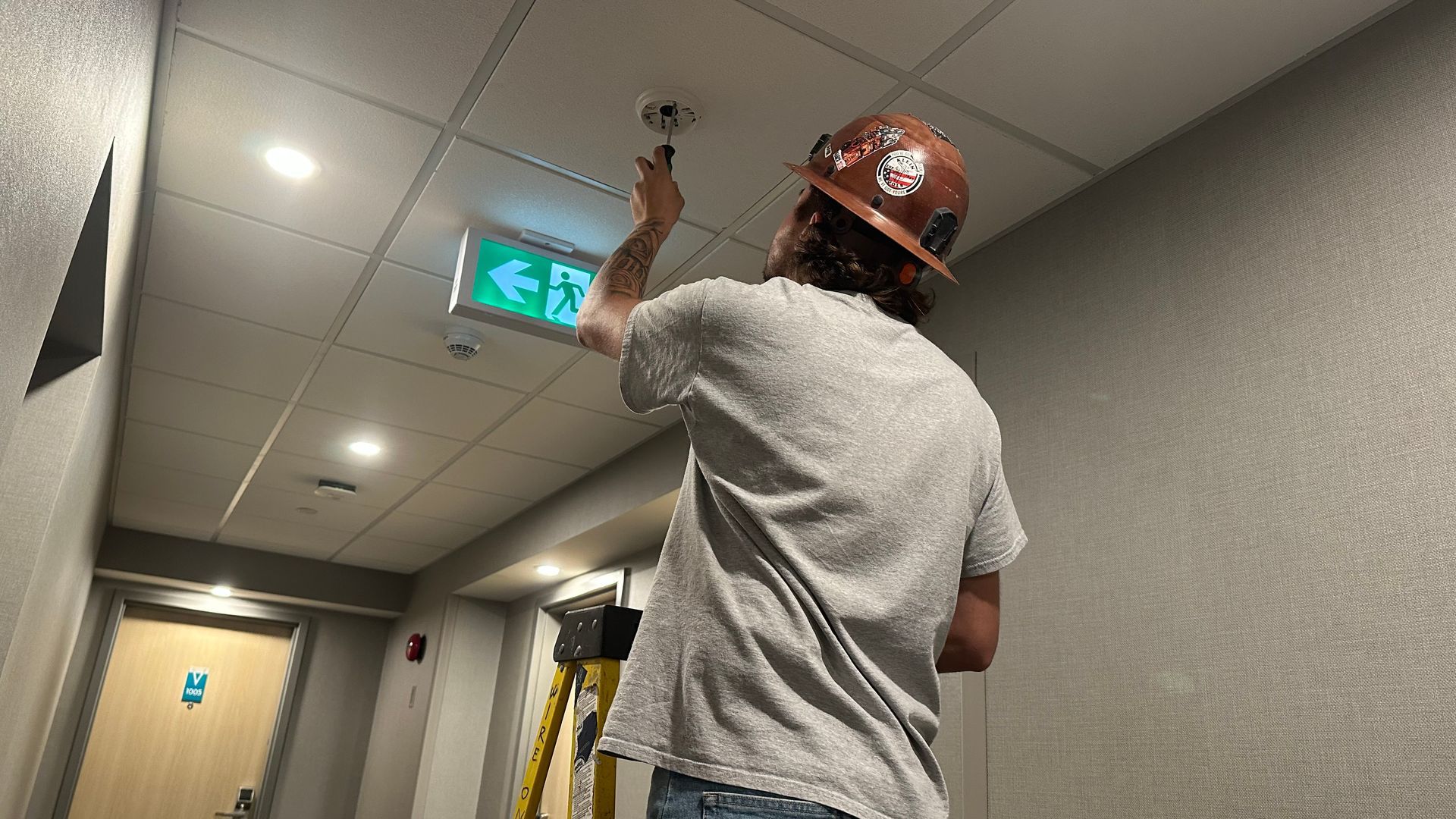 A person in a hard hat works on a ceiling fixture in a hallway, with an exit sign visible.