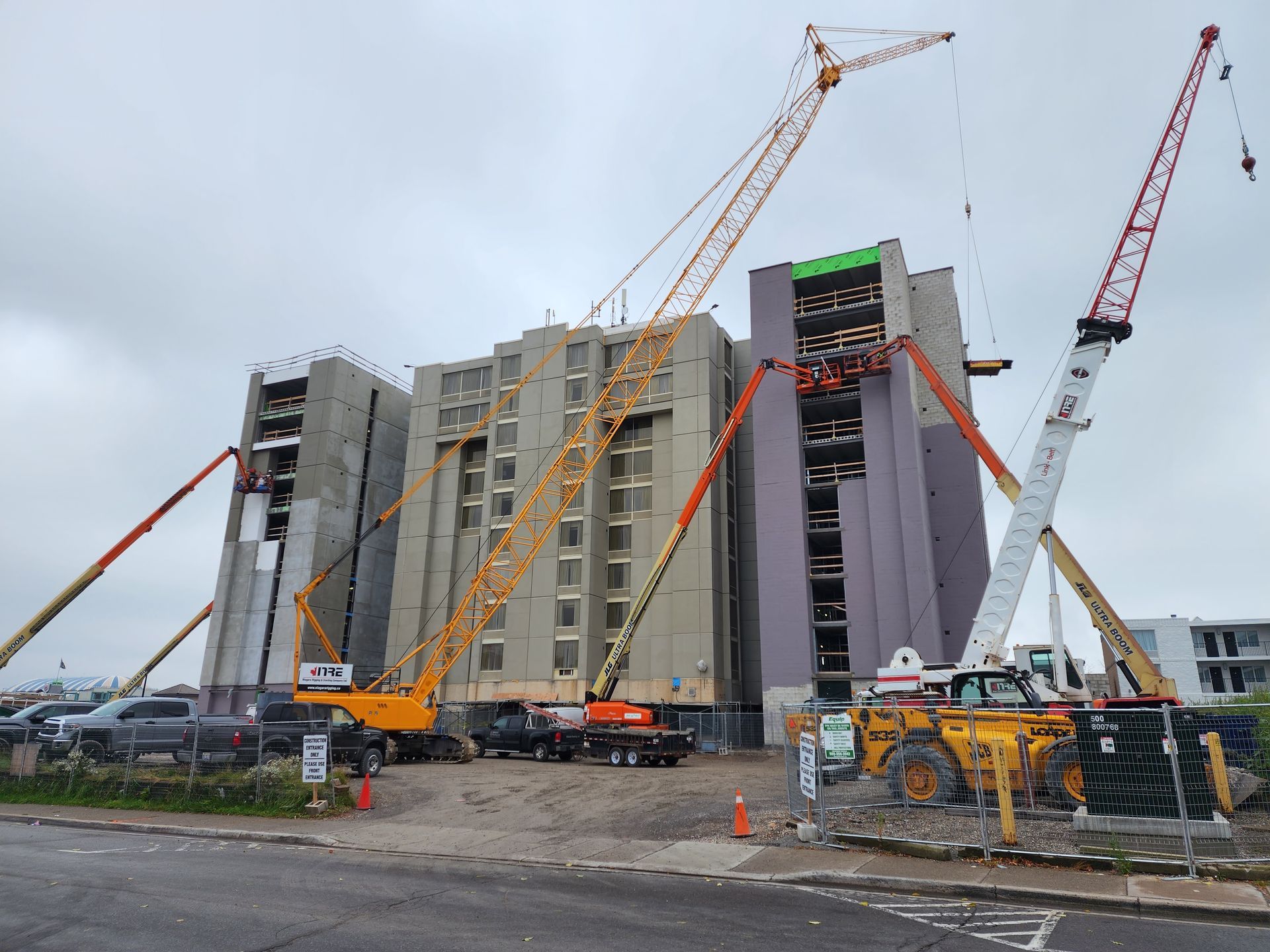 Construction site with several cranes surrounding a multi-story building under construction. Cranes are yellow, white, and red. Overcast sky.