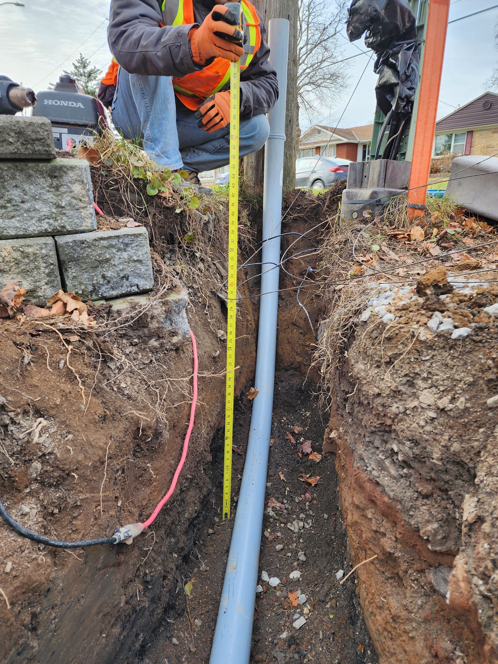 A person in work attire measures a gray pipe in a narrow trench with a yellow tape measure. Outdoor setting.