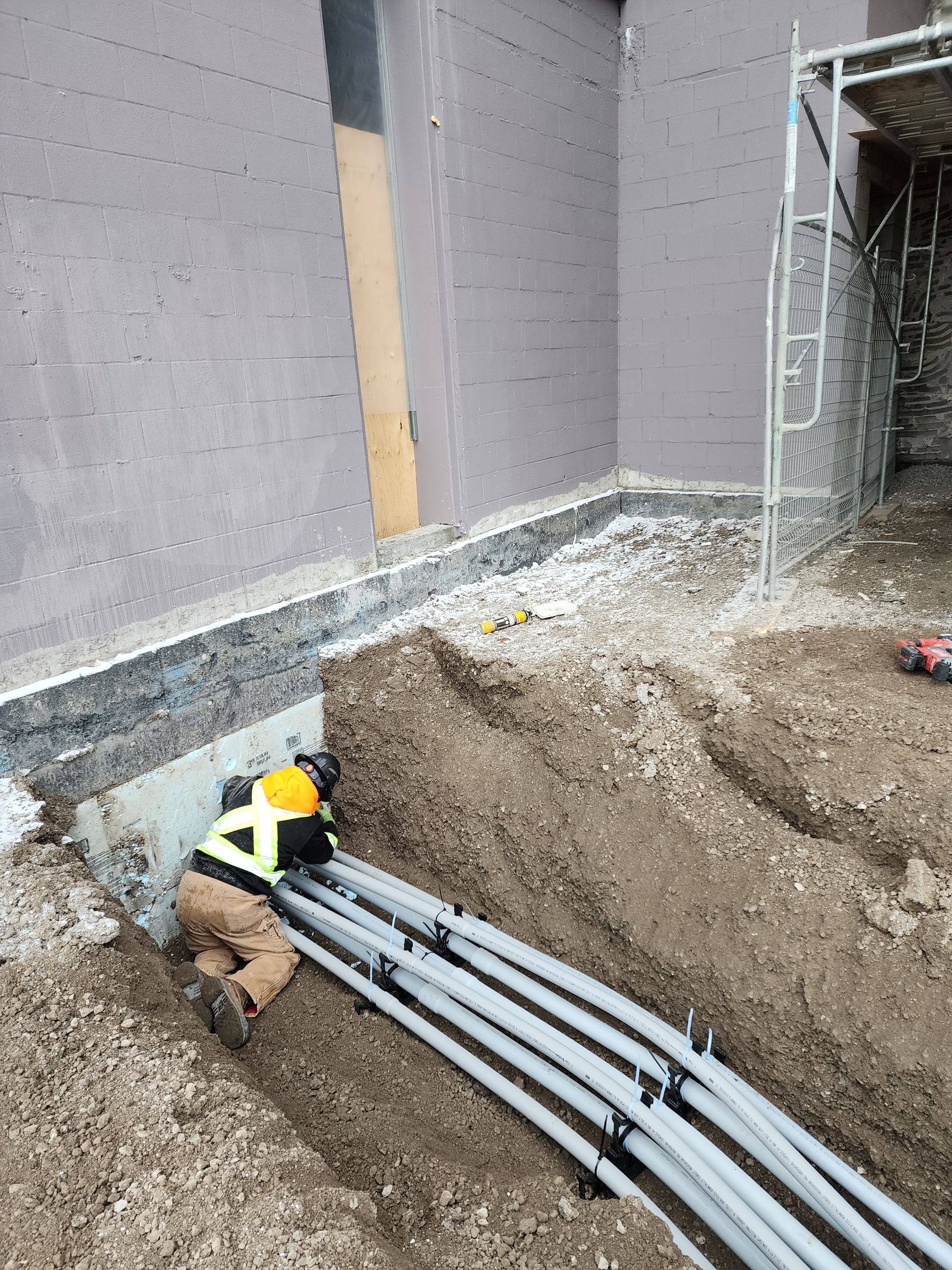 Construction worker installing gray conduit pipes in a trench next to a building wall. He is wearing a yellow vest and hard hat.