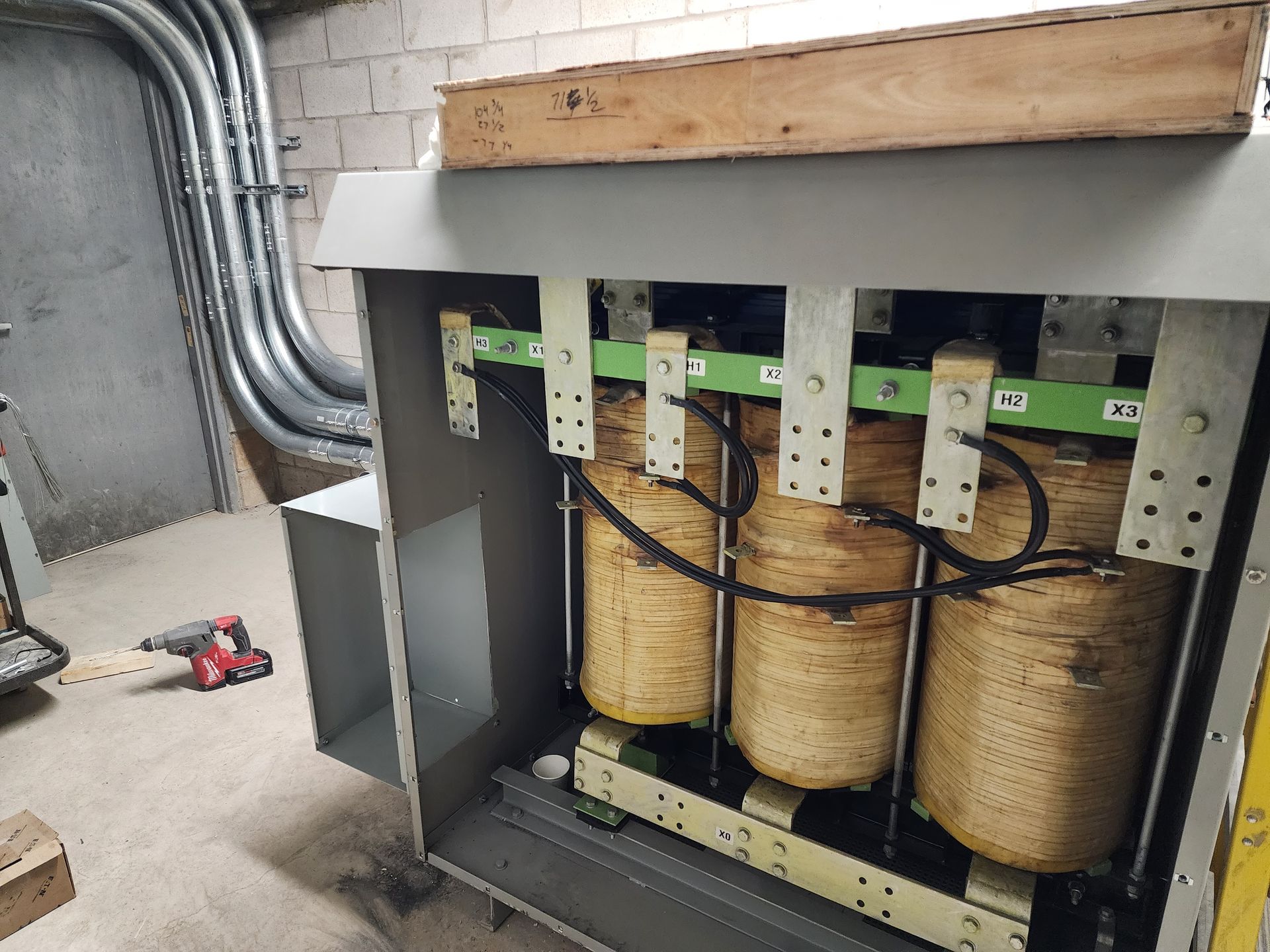 Three large electrical transformers in a metal enclosure, with exposed coils and wiring. A workshop setting with conduit and tools visible.
