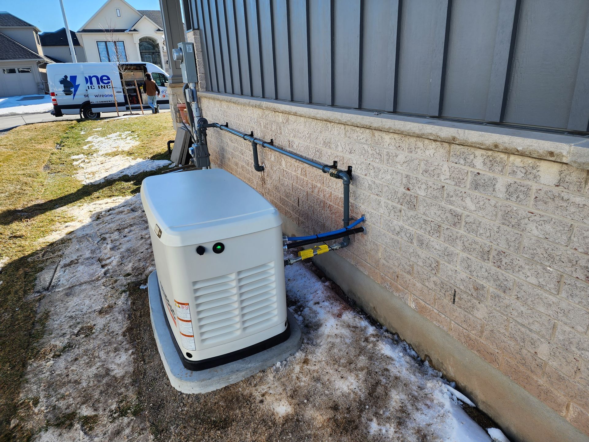A generator installed outside a house with snow on the ground. A metal pipe runs along the wall to it.