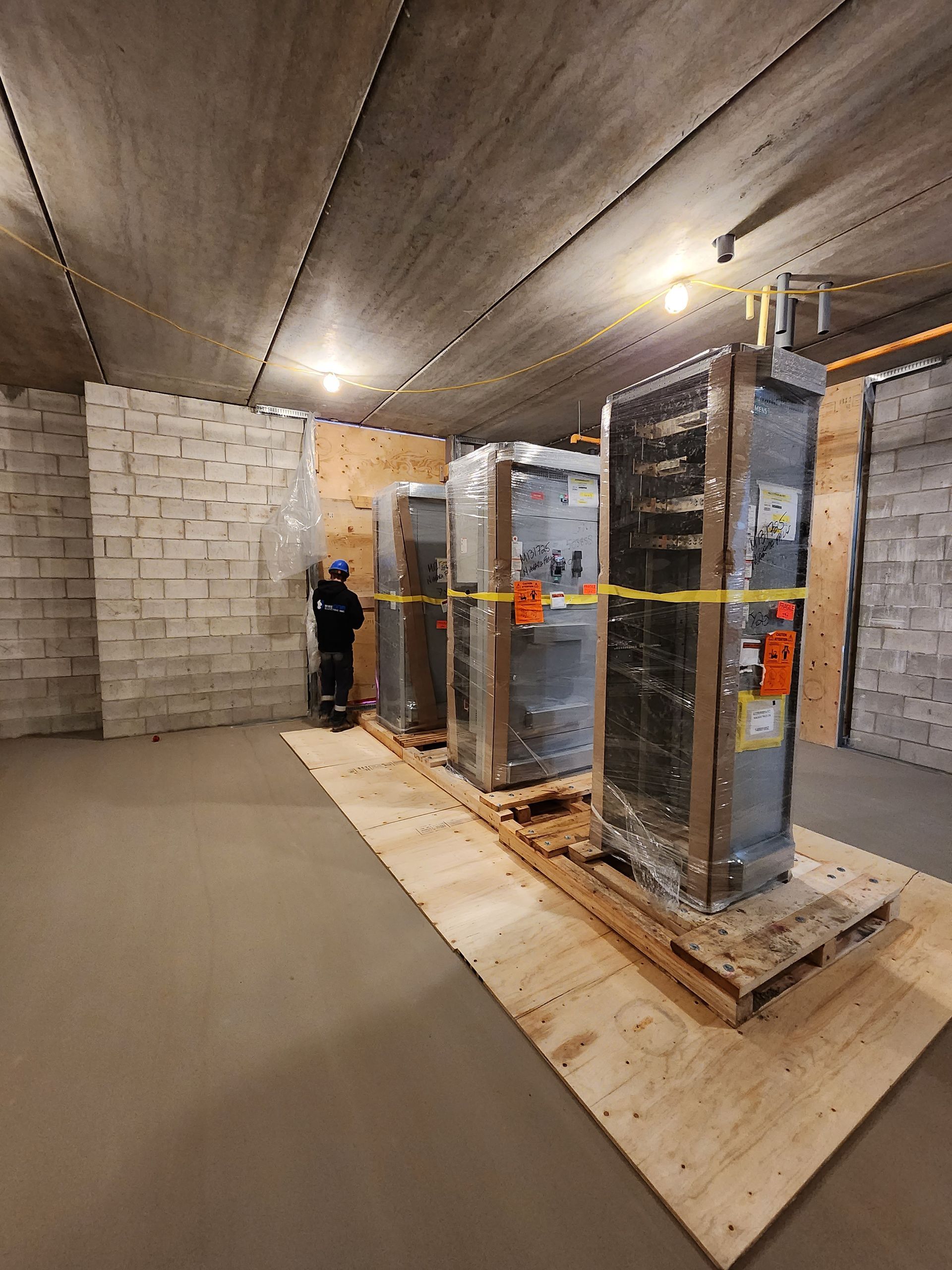 A person stands near three large electrical cabinets wrapped in plastic, on a wooden platform inside a room with concrete walls and ceiling.