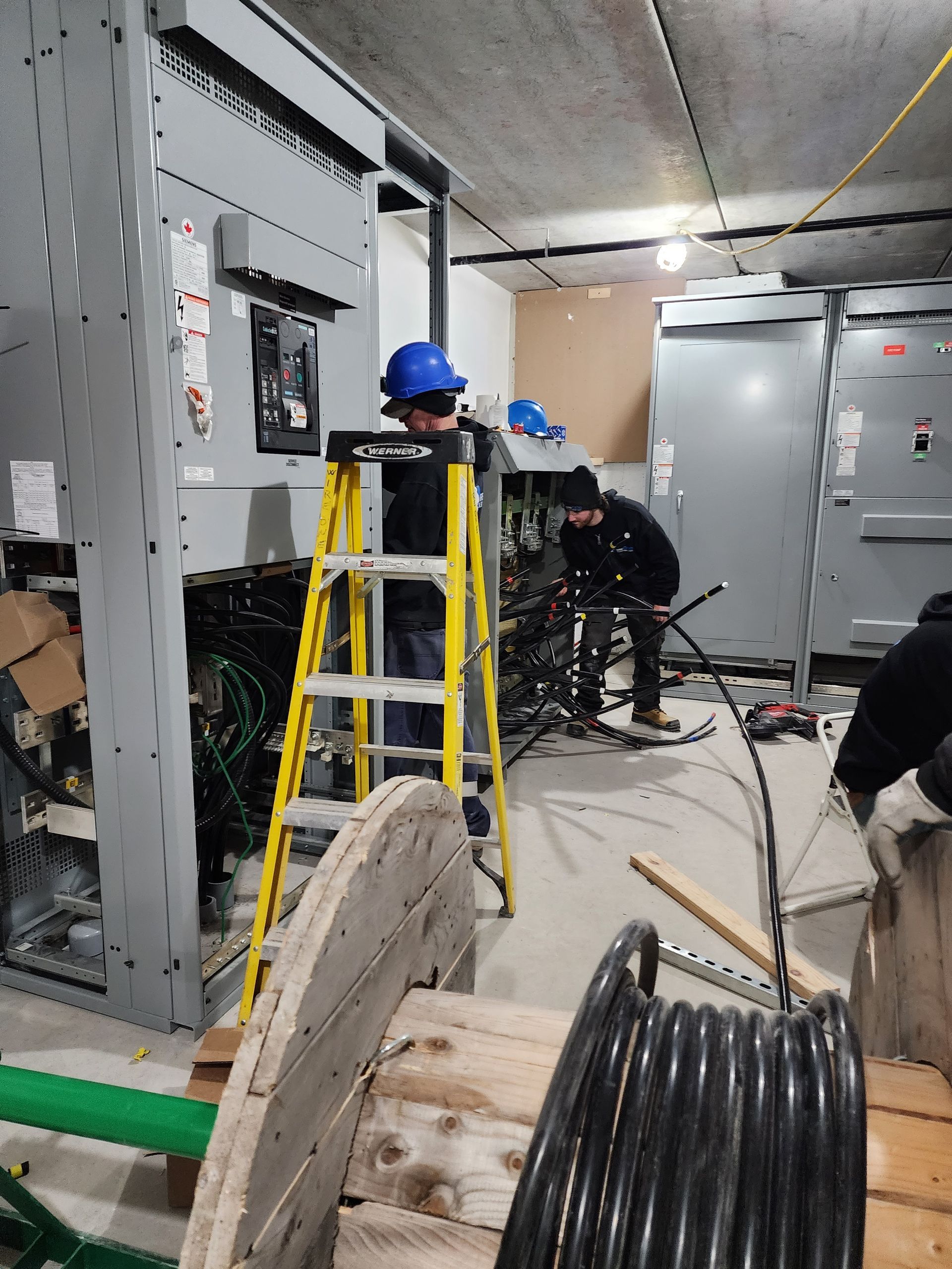 Two electricians work in an electrical room. One on a ladder works on a panel; another sorts wires. A large cable reel is in the foreground.