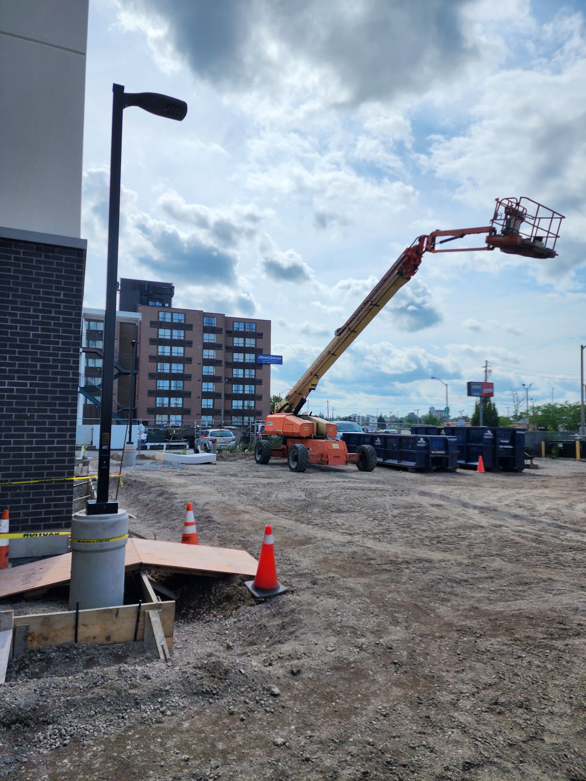 Construction site with an elevated work platform; a lamppost stands in the foreground. A multistory building and cloudy sky fill the background.