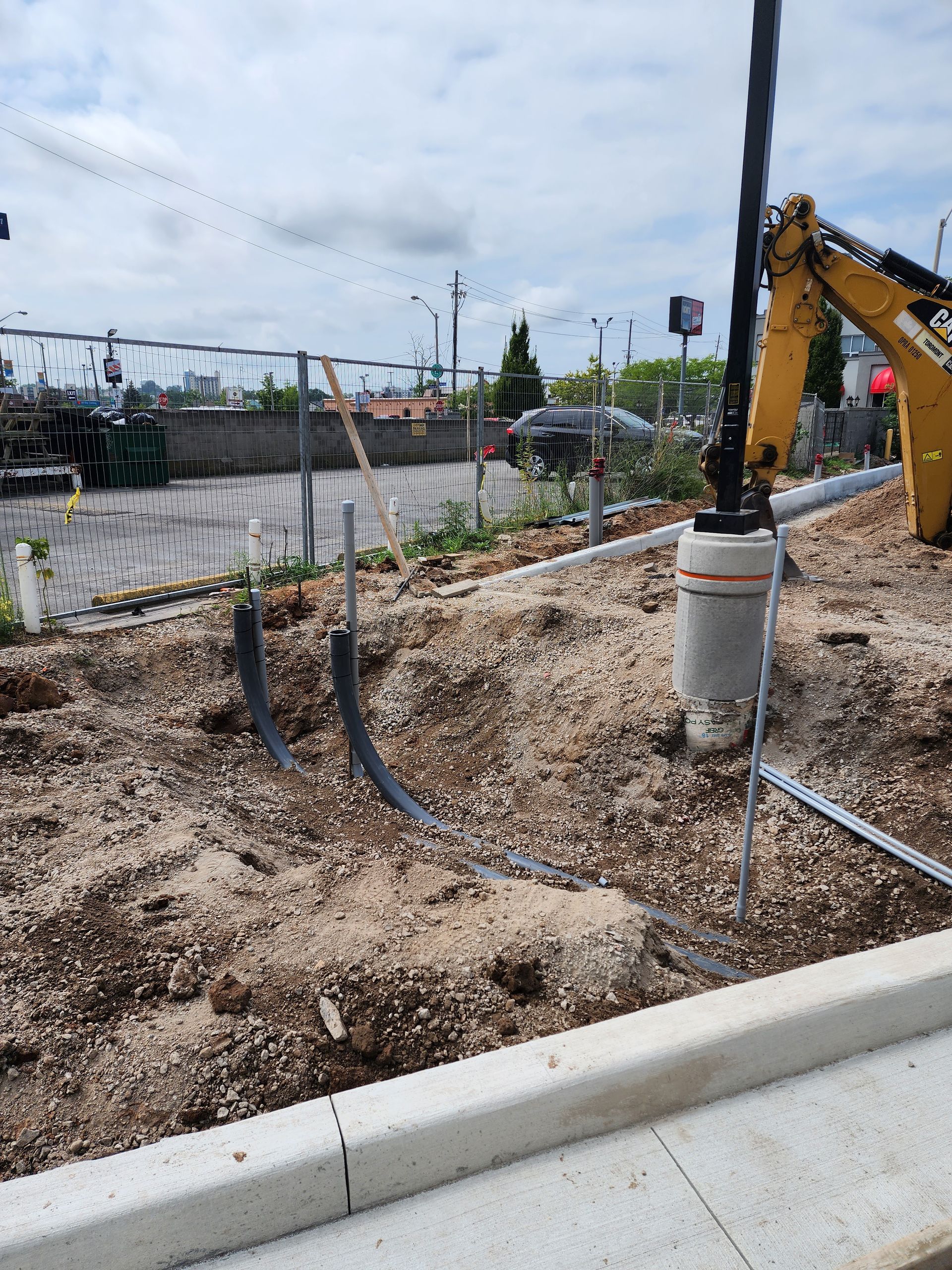 Construction site with exposed wiring and a concrete curb. A yellow excavator arm reaches toward a light pole.