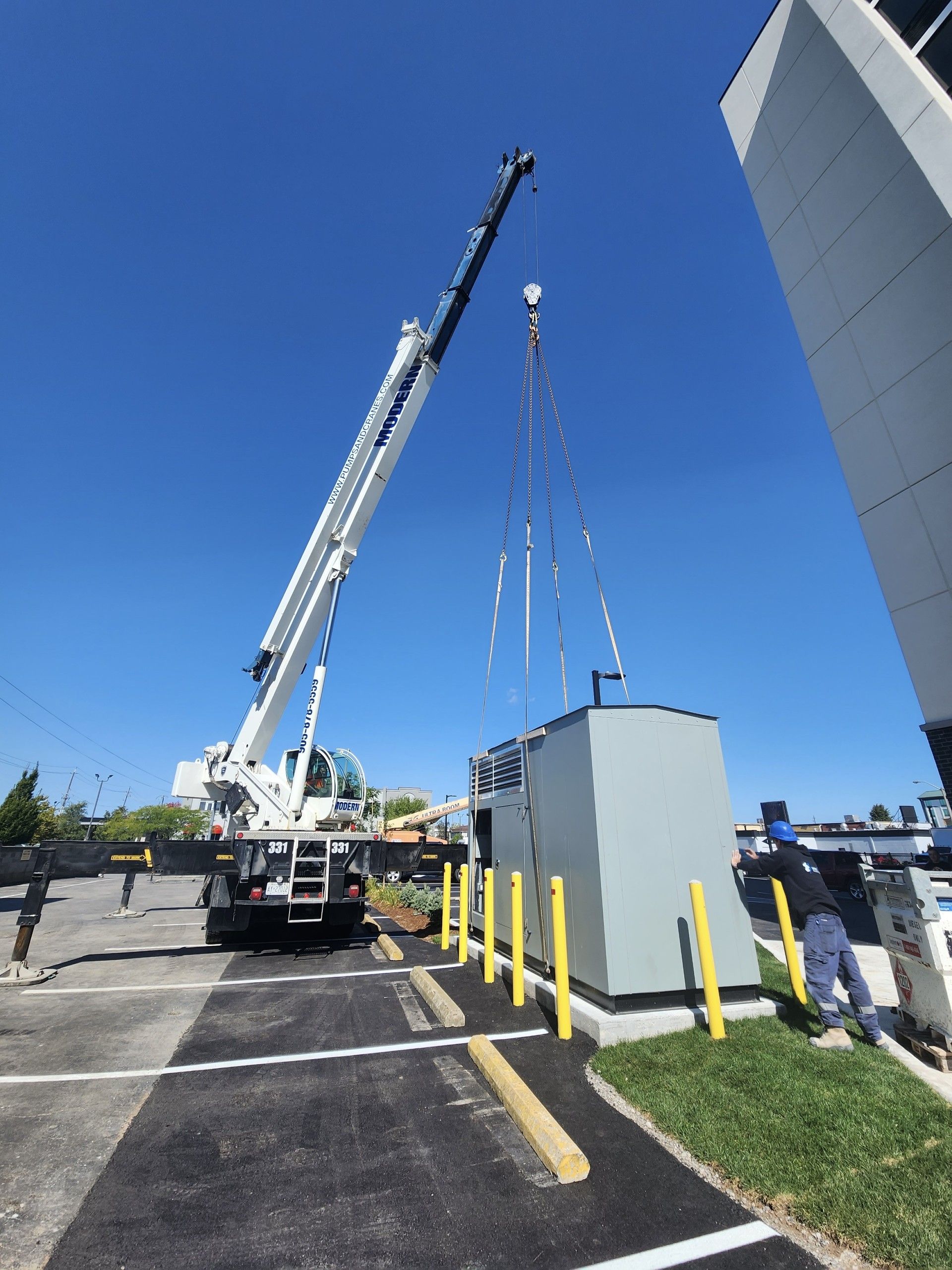 A large crane lifts a gray electrical unit. It's a sunny day with the crane beside a building on a paved lot.