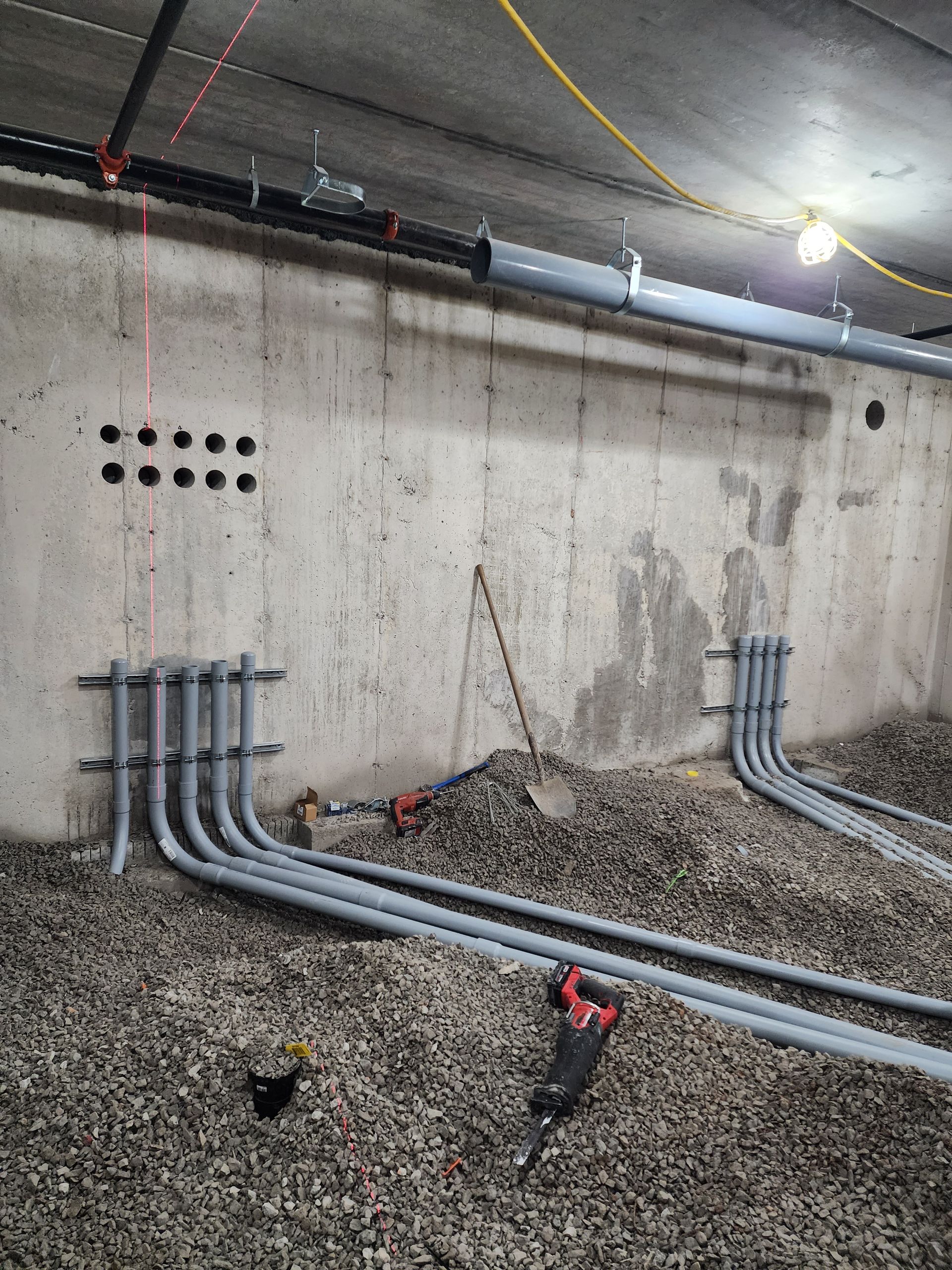 Gray pipes installed on a concrete wall with a scattered gravel floor, presumably under construction. A red laser level line is visible.