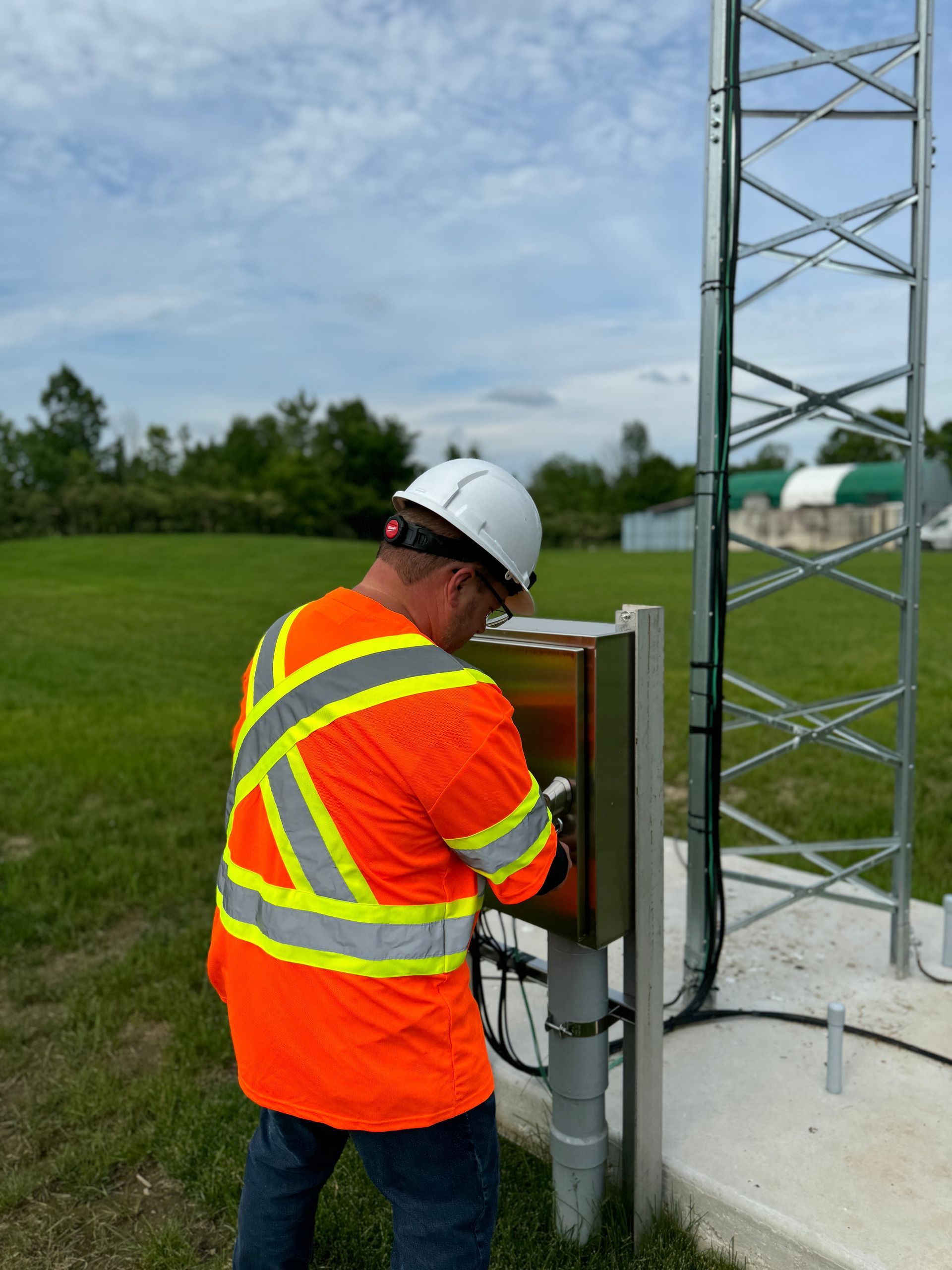 A person in an orange safety vest and hard hat working on an outdoor equipment box next to a tall communications tower.