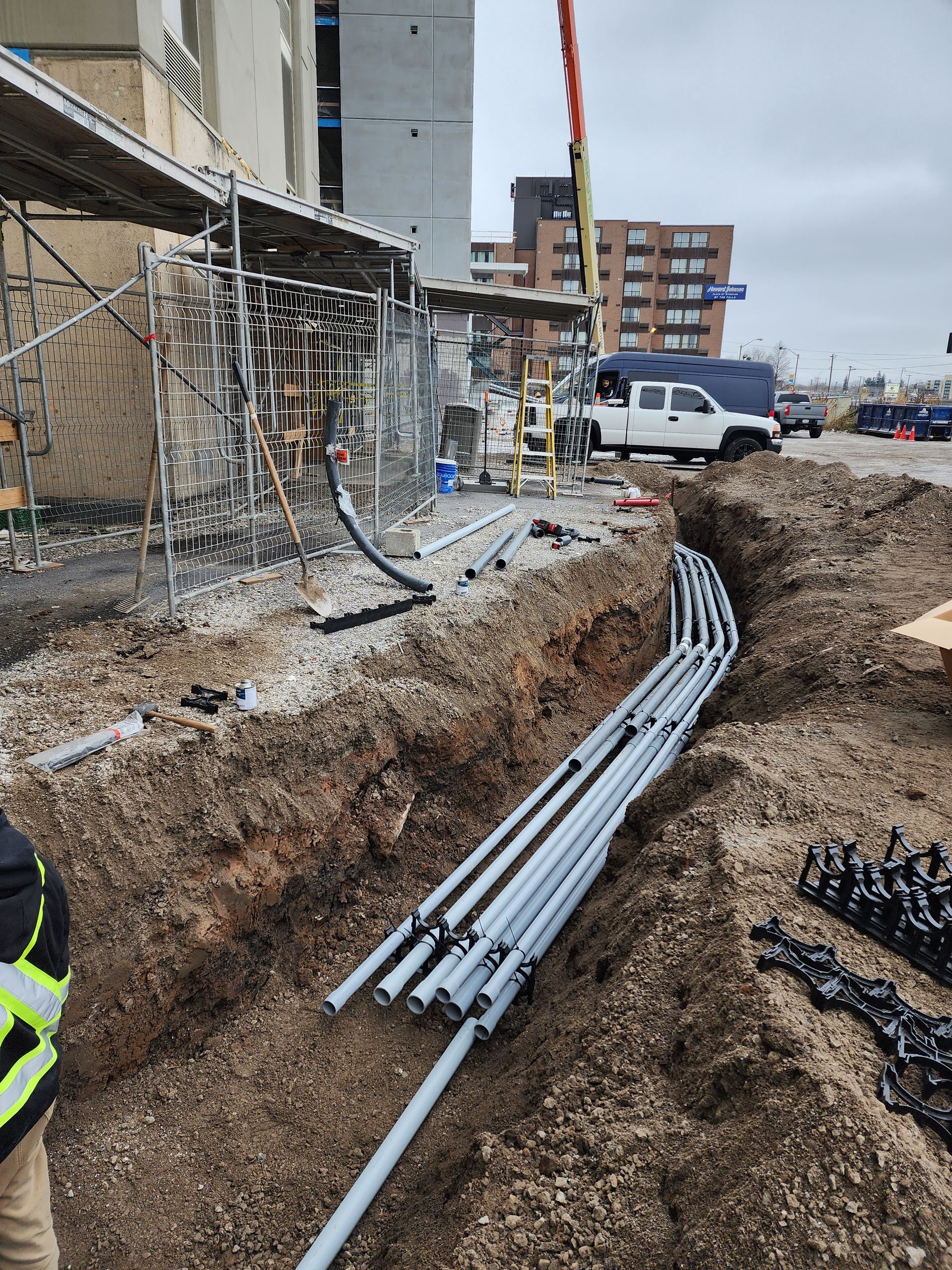 Construction site with a trench holding electrical conduits; scaffolding next to building under construction, a worker in safety gear.