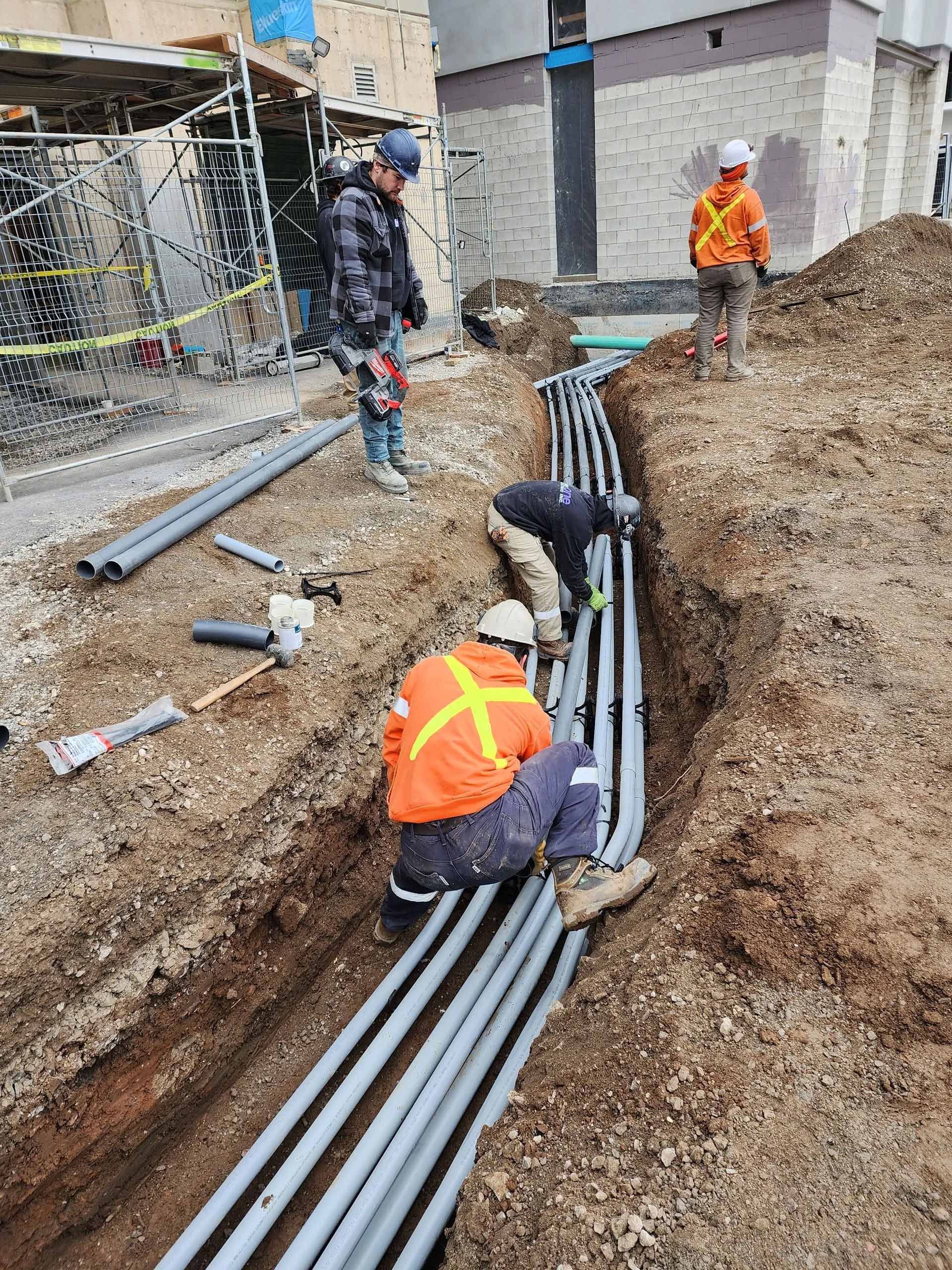 Construction workers installing gray conduit pipes in an open trench on a dirt construction site.
