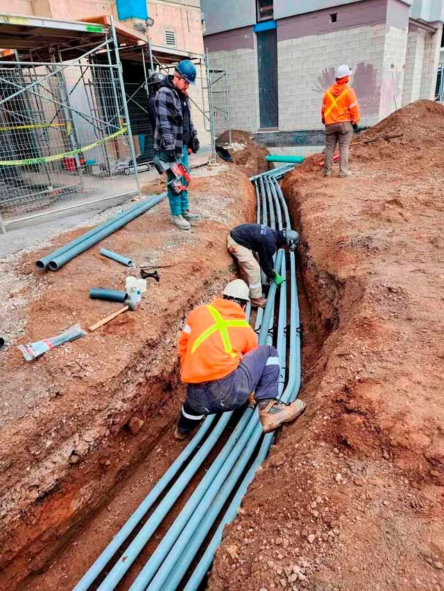 Construction workers installing gray conduit pipes in an open trench on a dirt construction site. One worker wears an orange vest.