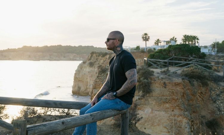 Man sitting on a wooden fence by a rocky seaside cliff at sunset