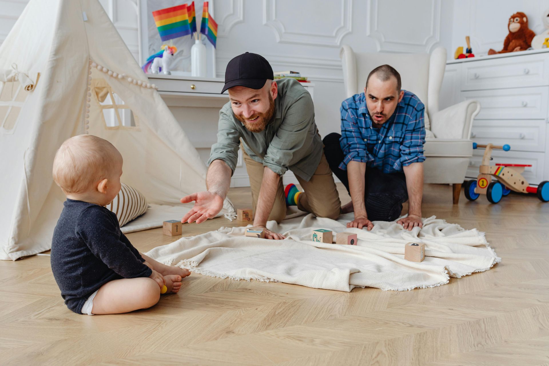 Two men playing with a baby on a rug in a playroom; tent, rainbow flag, and toys visible.