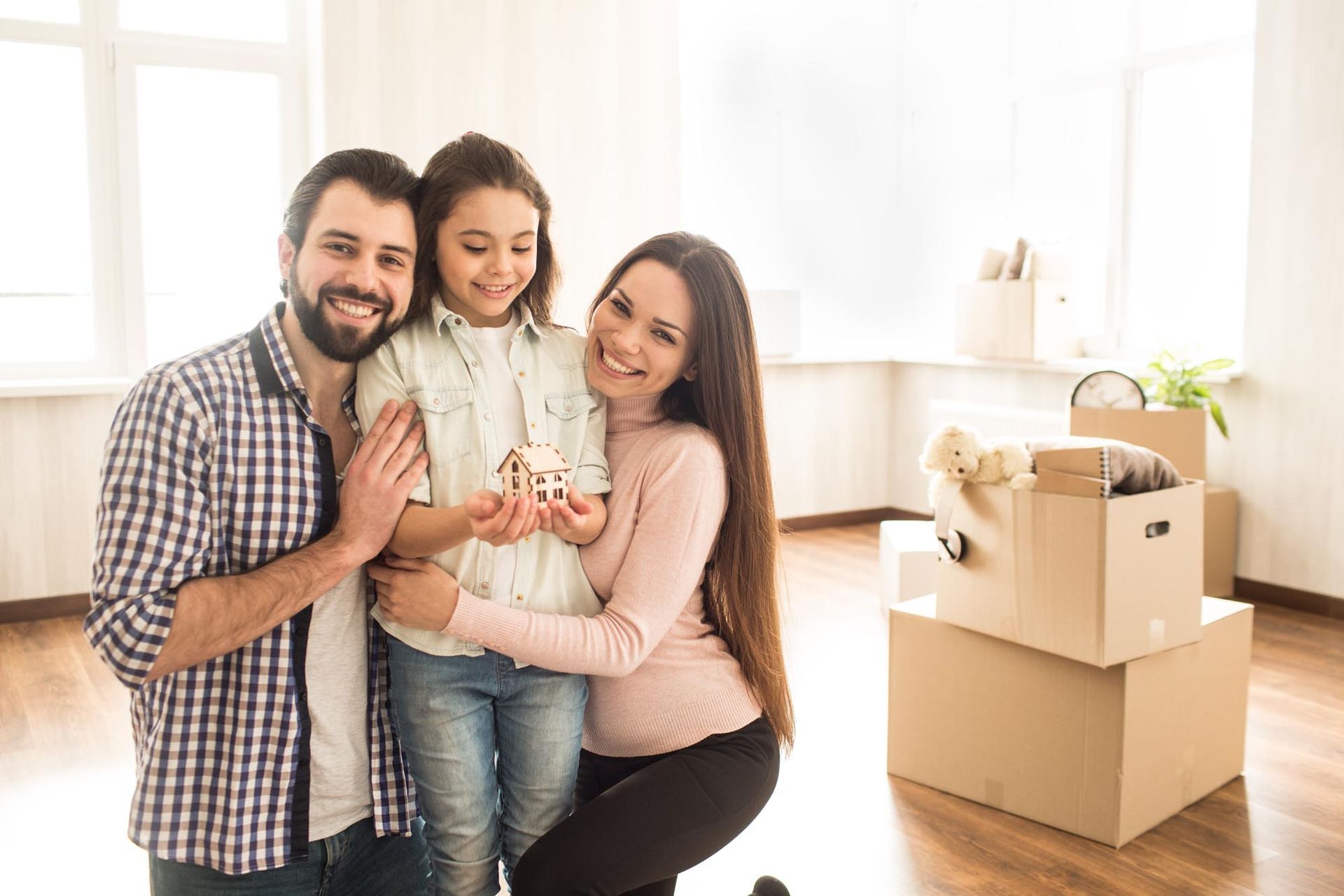 Family of three smiles, daughter holds a toy house in new home with moving boxes.