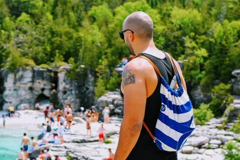 A man with a backpack is standing on a beach looking at the water.