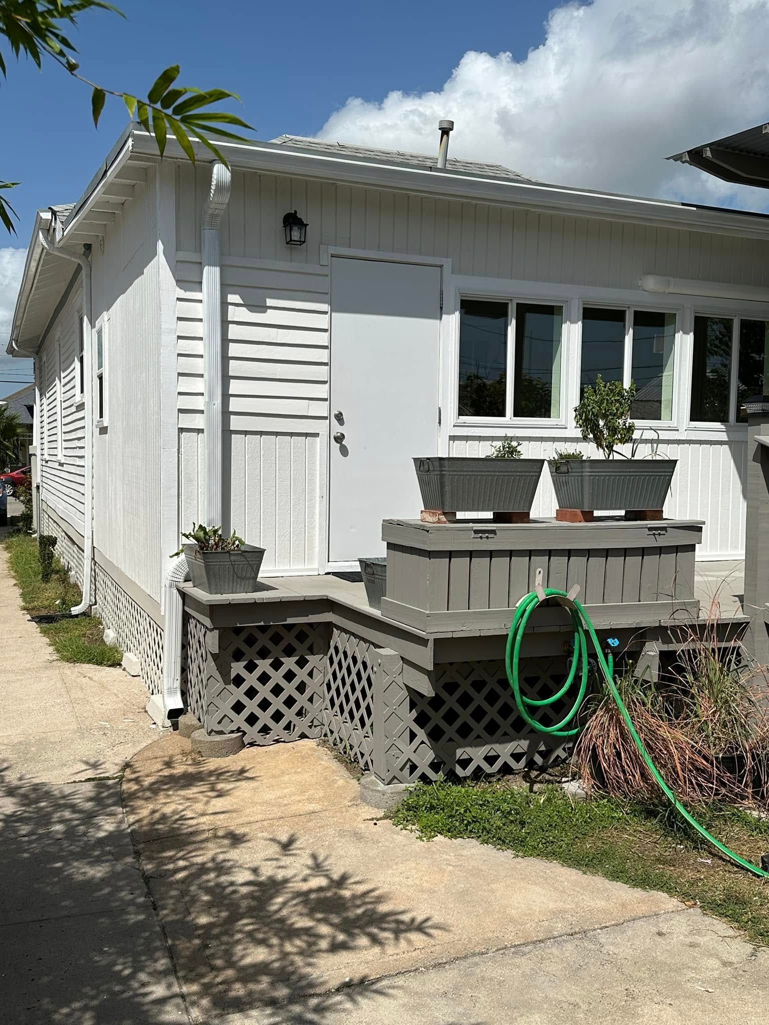 Small white house exterior with porch, windows, potted plants, and a green garden hose on the side.